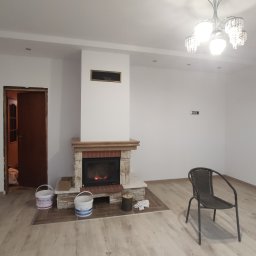 Azariasz Janicki ZIAJ-TECH - Jasne wnętrze z kominkiem obłożonym cegłą i drewnem, widoczne puszki z farbą, krzesło i parkiet. Świeżo pomalowane ściany i sufit z oświetleniem punktowym.