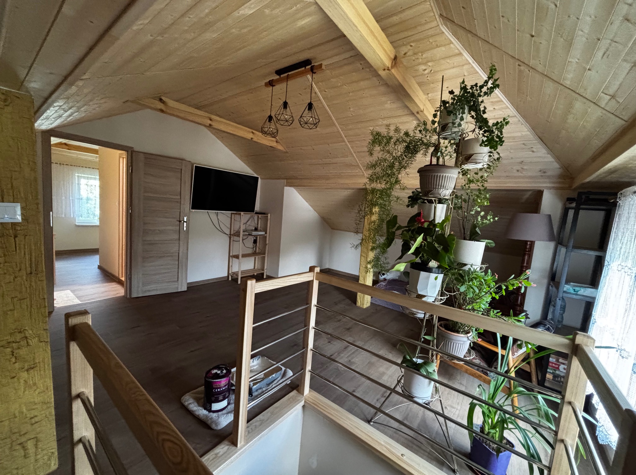 Drewniane wykończenie poddasza z widocznymi belkami, oświetleniem i roślinami doniczkowymi. Na podłodze puszka farby, co sugeruje prace wykończeniowe.