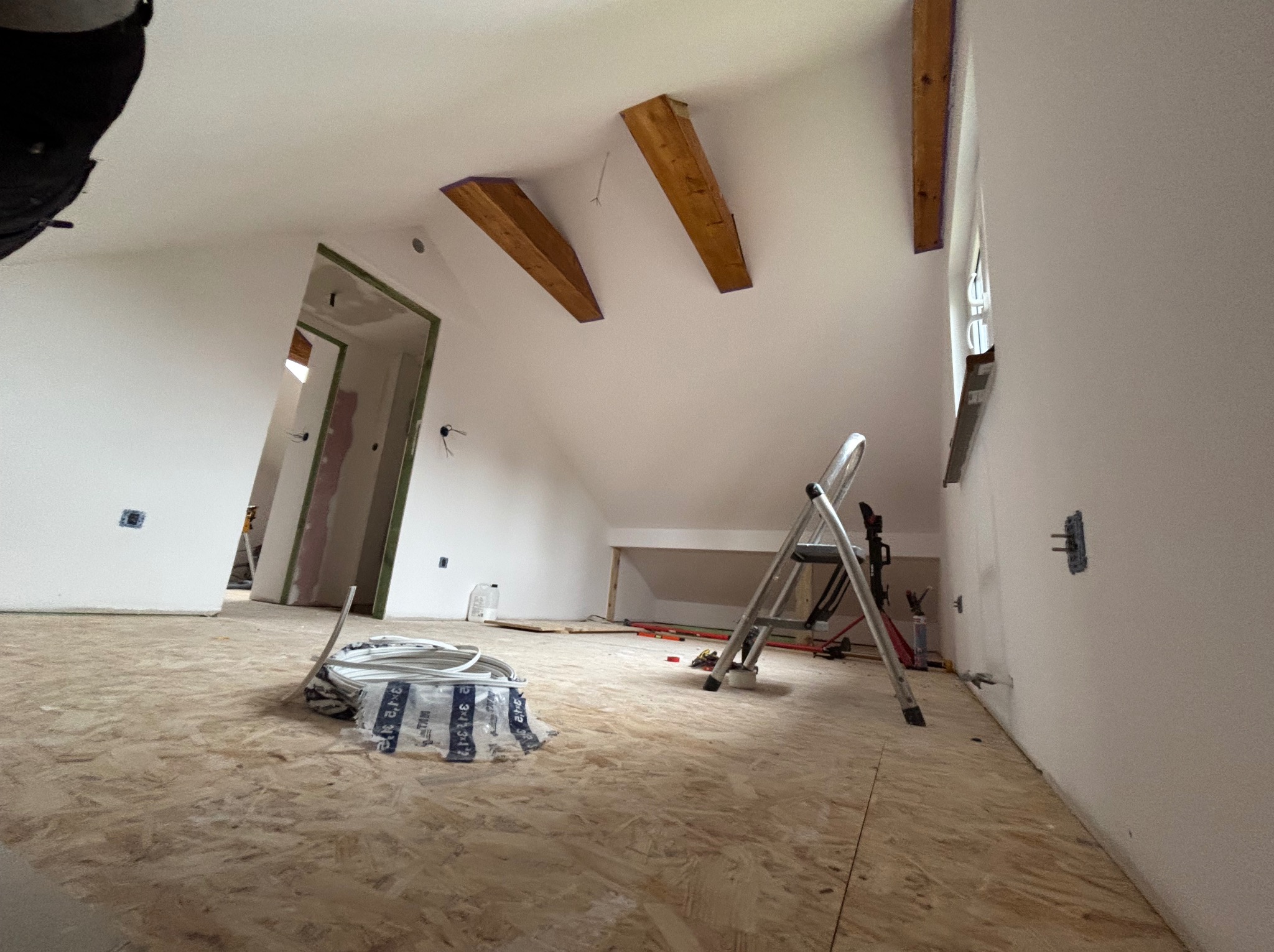 Poddasze w trakcie wykończenia: surowe drewniane belki, białe ściany, płyta OSB na podłodze, drabina i przewody elektryczne. Widoczne gniazdka elektryczne.