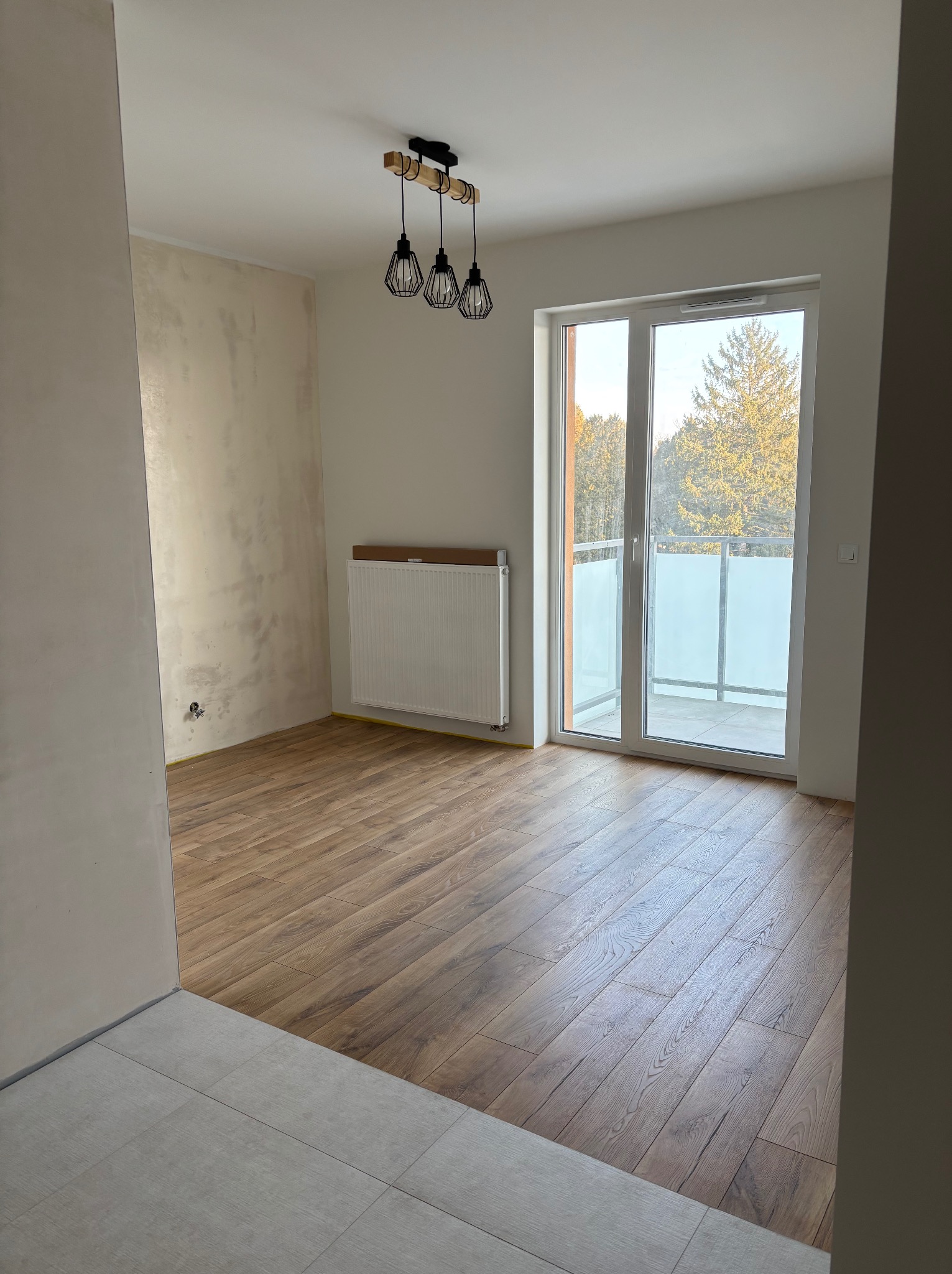 Jasne wnętrze po remoncie: parkiet, ściana z fakturą, balkon z widokiem na drzewa. Nowoczesna lampa wisząca. Grzejnik pod oknem balkonowym.