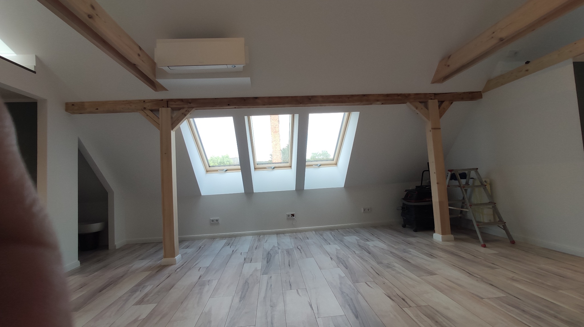 Jasne poddasze po remoncie z drewnianymi belkami, trzema oknami dachowymi i panelami podłogowymi imitującymi drewno. Widoczna drabina i odkurzacz.