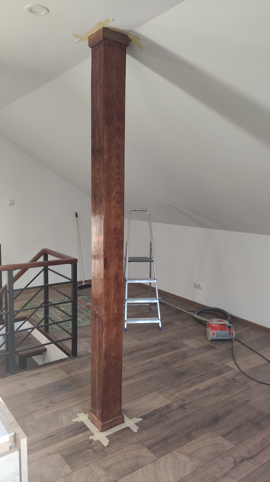 Drewniana kolumna po renowacji, zabezpieczona taśmą malarską u podstawy i góry. W tle drabina, odkurzacz i drewniana podłoga w jasnym, odnowionym wnętrzu poddasza.