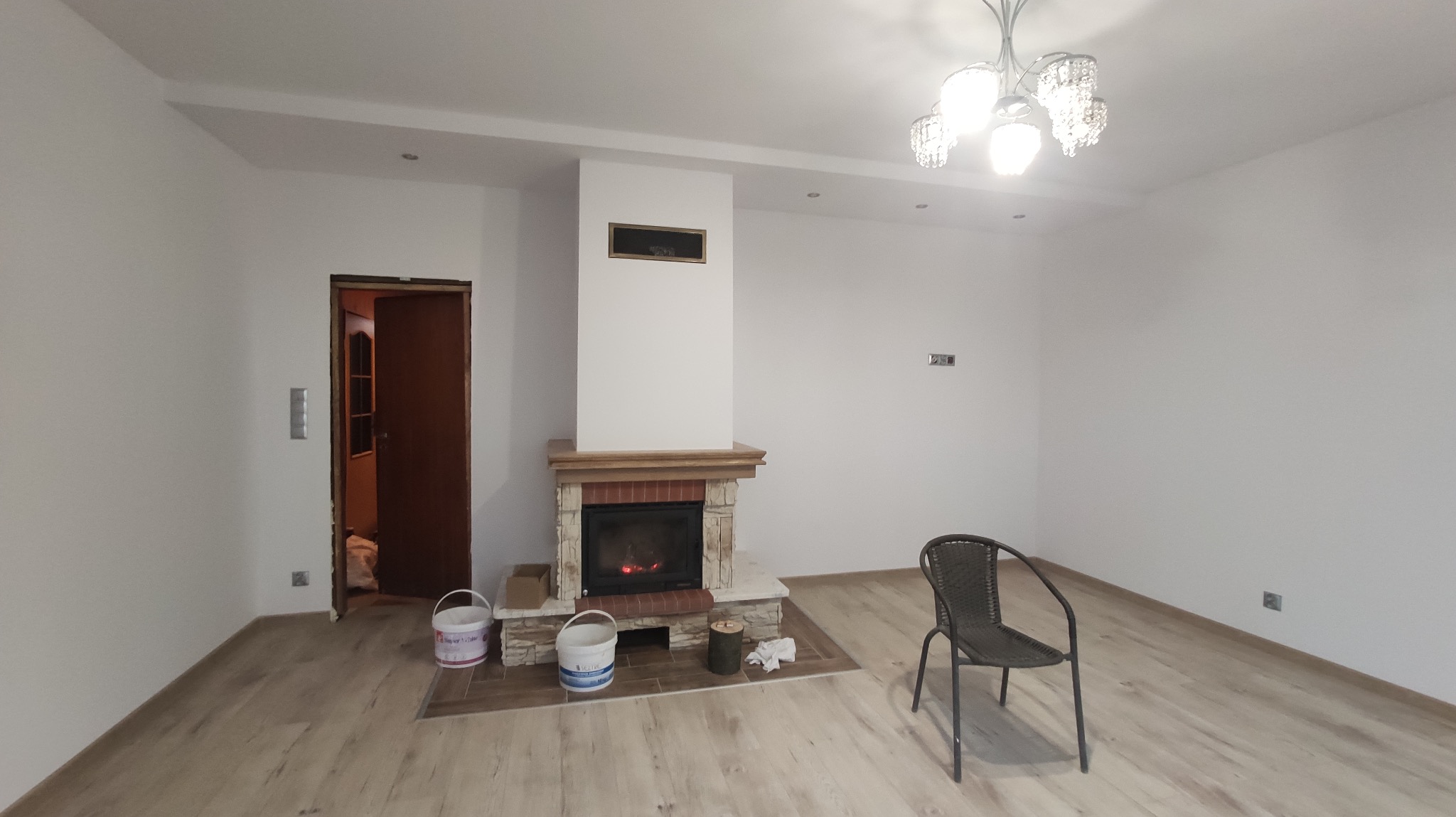 Jasne wnętrze z kominkiem obłożonym cegłą i drewnem, widoczne puszki z farbą, krzesło i parkiet. Świeżo pomalowane ściany i sufit z oświetleniem punktowym.