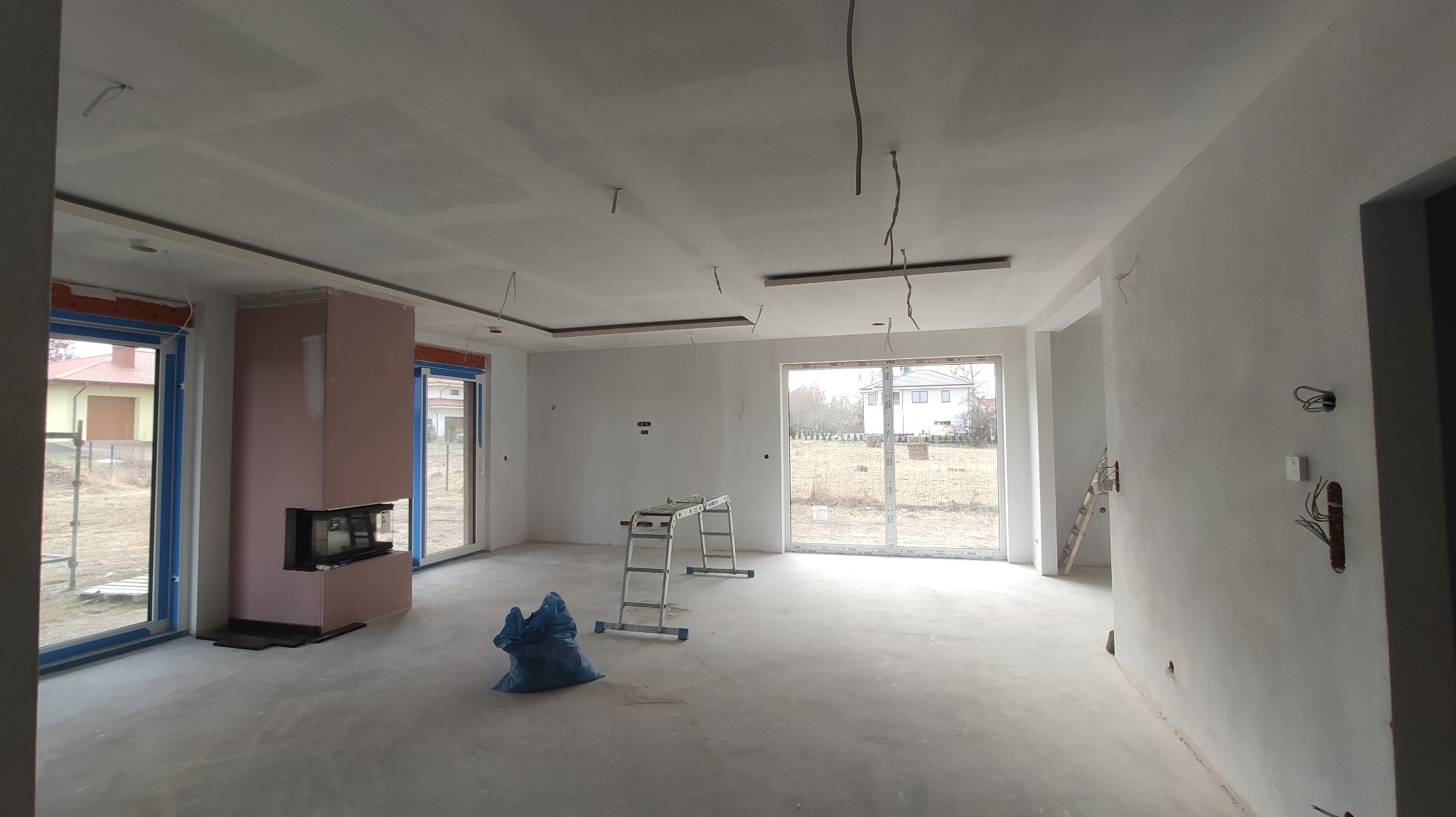 Wnętrze w trakcie wykończenia: sufit z płyt karton-gips, przygotowany kominek obudowany płytami, okna PCV, drabina i worki na podłodze. Widoczne kable i instalacje.