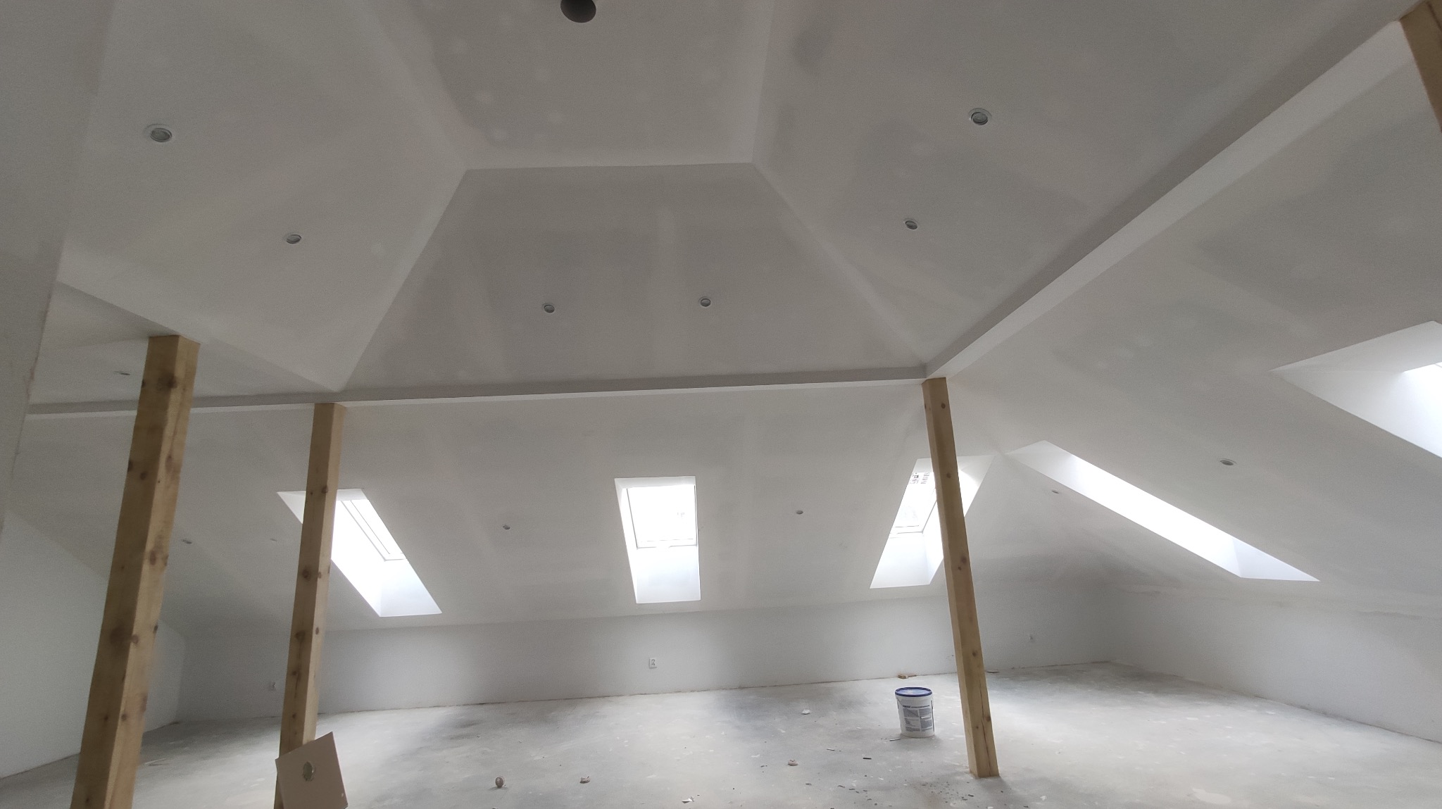 Poddasze po montażu płyt karton-gips z widocznymi oknami dachowymi i drewnianymi podporami. Sufit z wbudowanym oświetleniem punktowym. Ściany przygotowane do malowania.