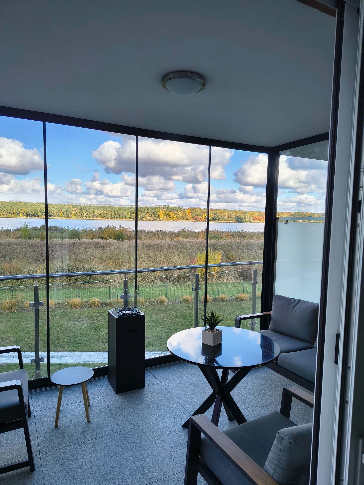 Zadaszony taras z widokiem na jezioro przez duże przesuwne okna. Nowoczesne meble ogrodowe i dekoracyjny kominek tworzą przytulną przestrzeń.