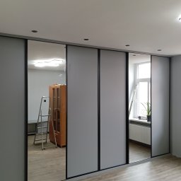 Katarzyna Mamorska - Nowoczesna szafa przesuwna z szarymi frontami i czarnymi obramowaniami, częściowo z lustrami, odbijającymi wnętrze pokoju z drabiną i szafką.