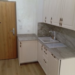 Katarzyna Mamorska - Jasna zabudowa kuchenna z marmurowym blatem i złotymi uchwytami, widoczna obok drewnianych drzwi wejściowych. Minimalistyczny design, eleganckie wykończenie.