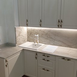 Katarzyna Mamorska - Elegancka kuchnia na wymiar w Pasieczniku z białymi frontami, marmurowym blatem i złotymi uchwytami. Widoczny zlew i płyta indukcyjna, oświetlenie LED pod szafkami.