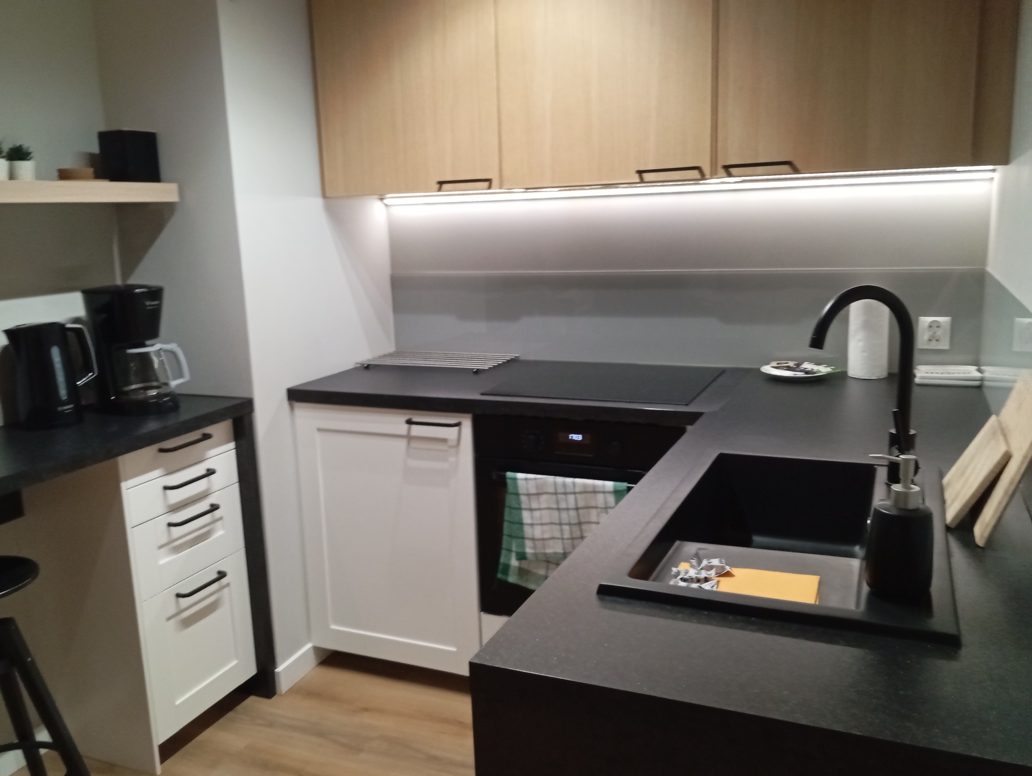 Nowoczesna kuchnia z drewnianymi szafkami, czarnym blatem i białymi frontami. Widoczny zlew z czarną armaturą i oświetlenie LED pod szafkami.