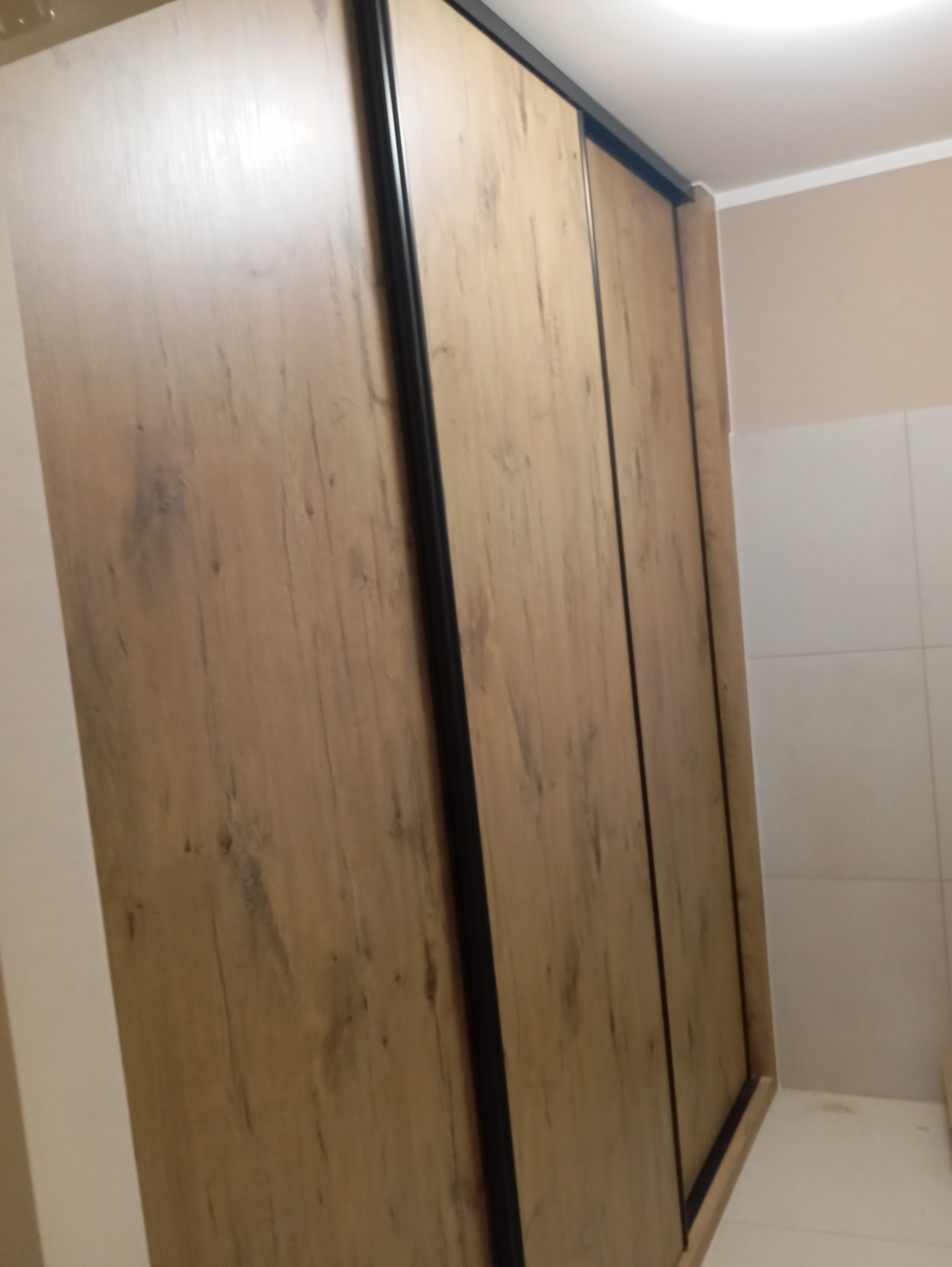Drewniana szafa przesuwna z czarnymi obramowaniami, widoczna z boku, z teksturą imitującą naturalne drewno. Minimalistyczny design w jasnym wnętrzu.