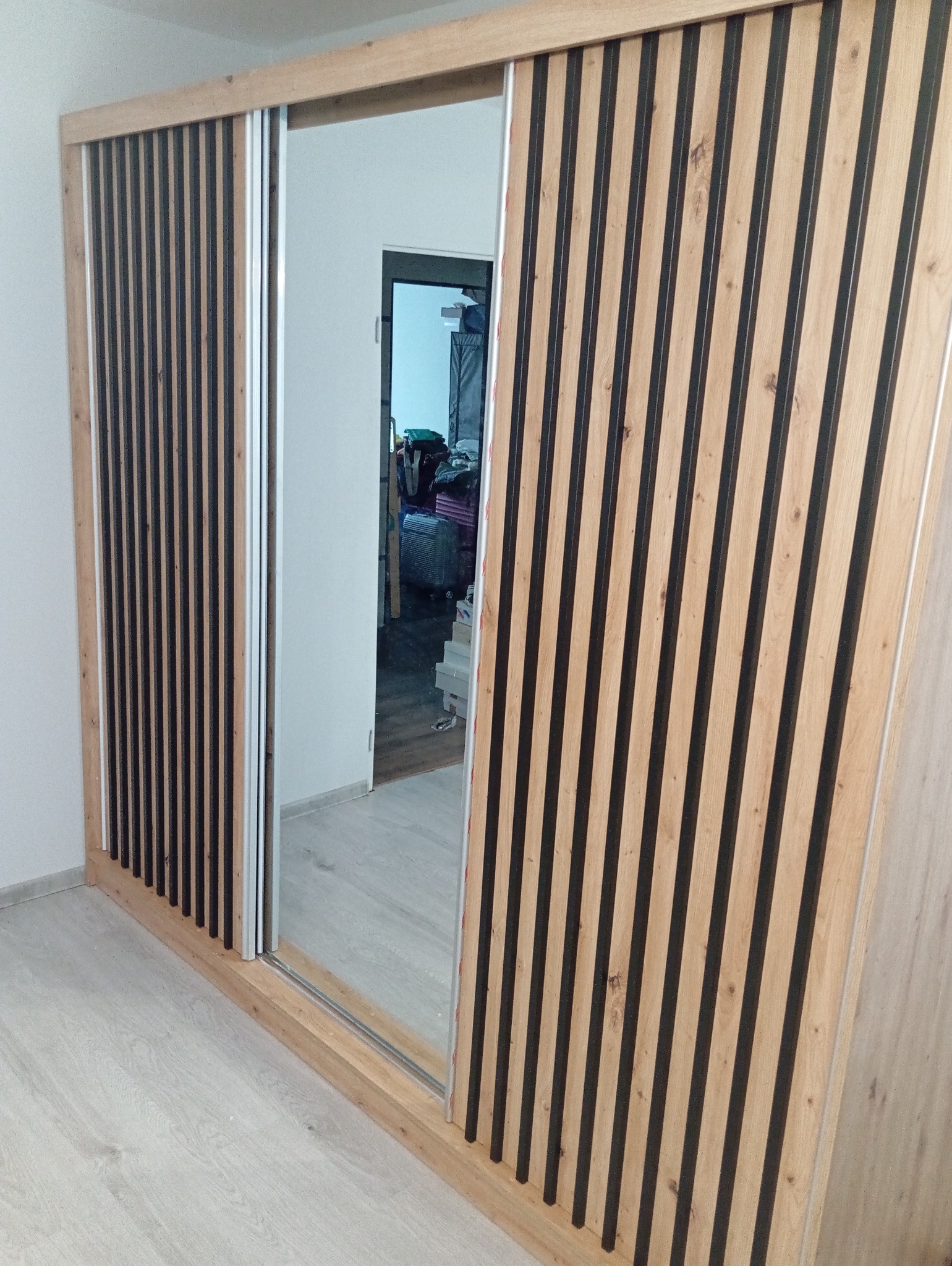 Nowoczesna szafa przesuwna z lustrem i dekoracyjnymi lamelami w odcieniach drewna i czerni. Widoczne detale wykończenia i system przesuwny.