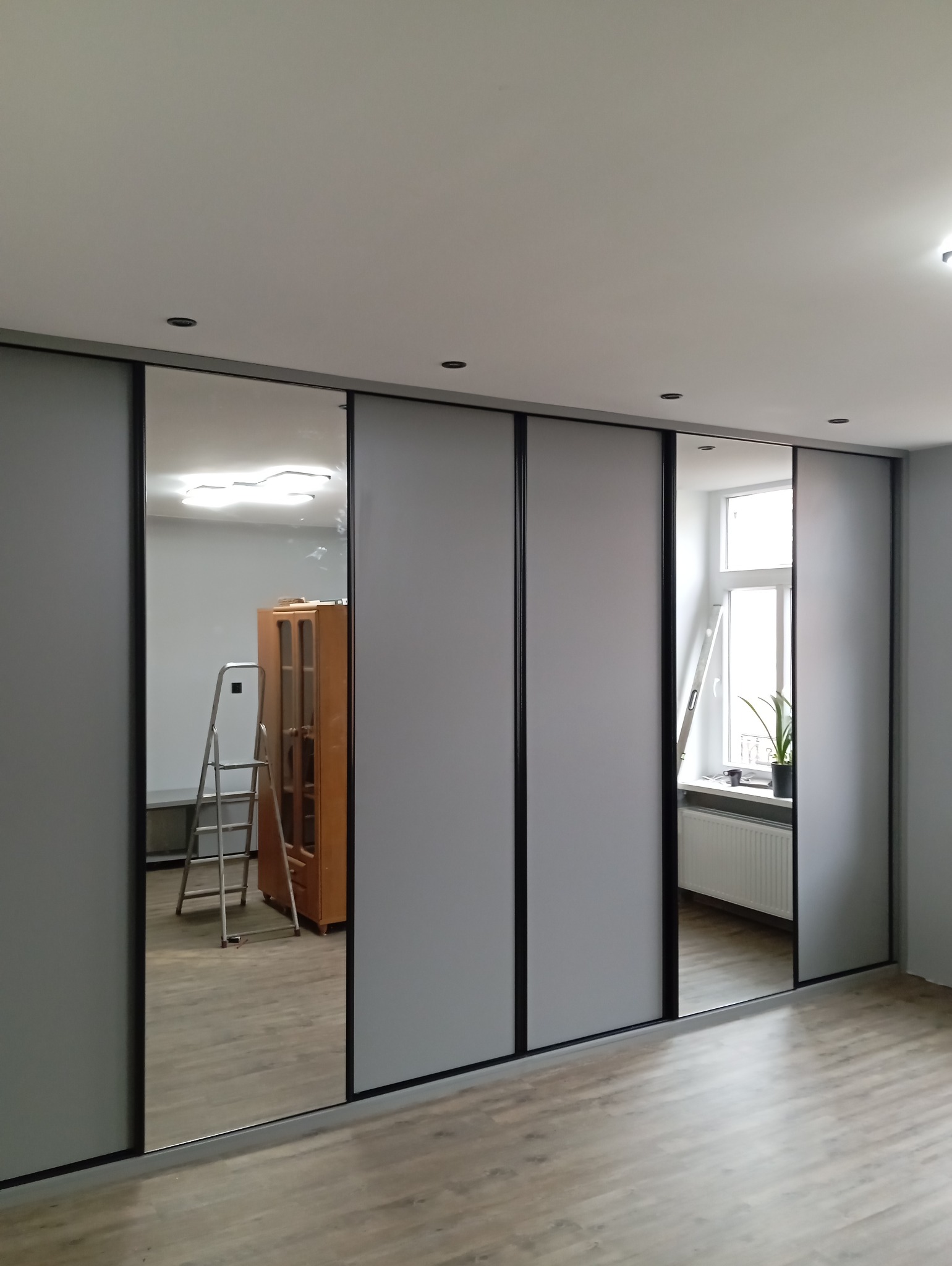 Nowoczesna szafa przesuwna z szarymi frontami i czarnymi obramowaniami, częściowo z lustrami, odbijającymi wnętrze pokoju z drabiną i szafką.