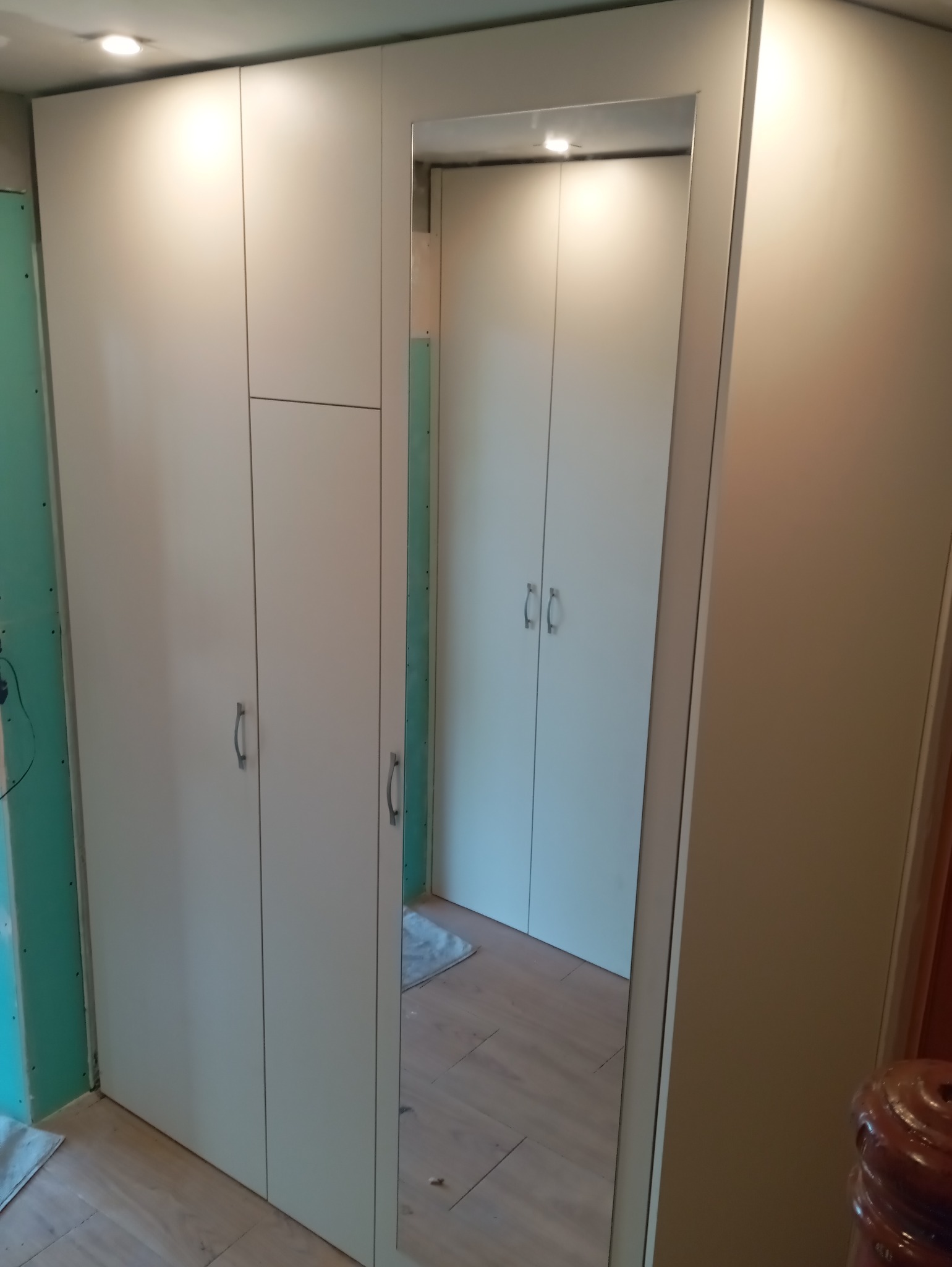 Biała szafa wnękowa z lustrem, widoczna w trakcie montażu. Ściana z płyty gipsowej w tle. Minimalistyczny design, srebrne uchwyty, oświetlenie punktowe.
