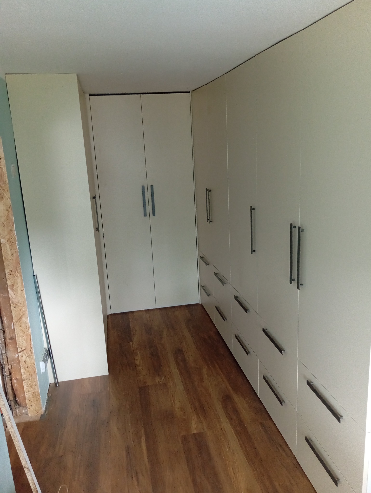 Jasna zabudowa szaf wnękowych z szufladami i długimi uchwytami, na podłodze panele imitujące drewno. Widoczny fragment ściany w trakcie wykończenia.