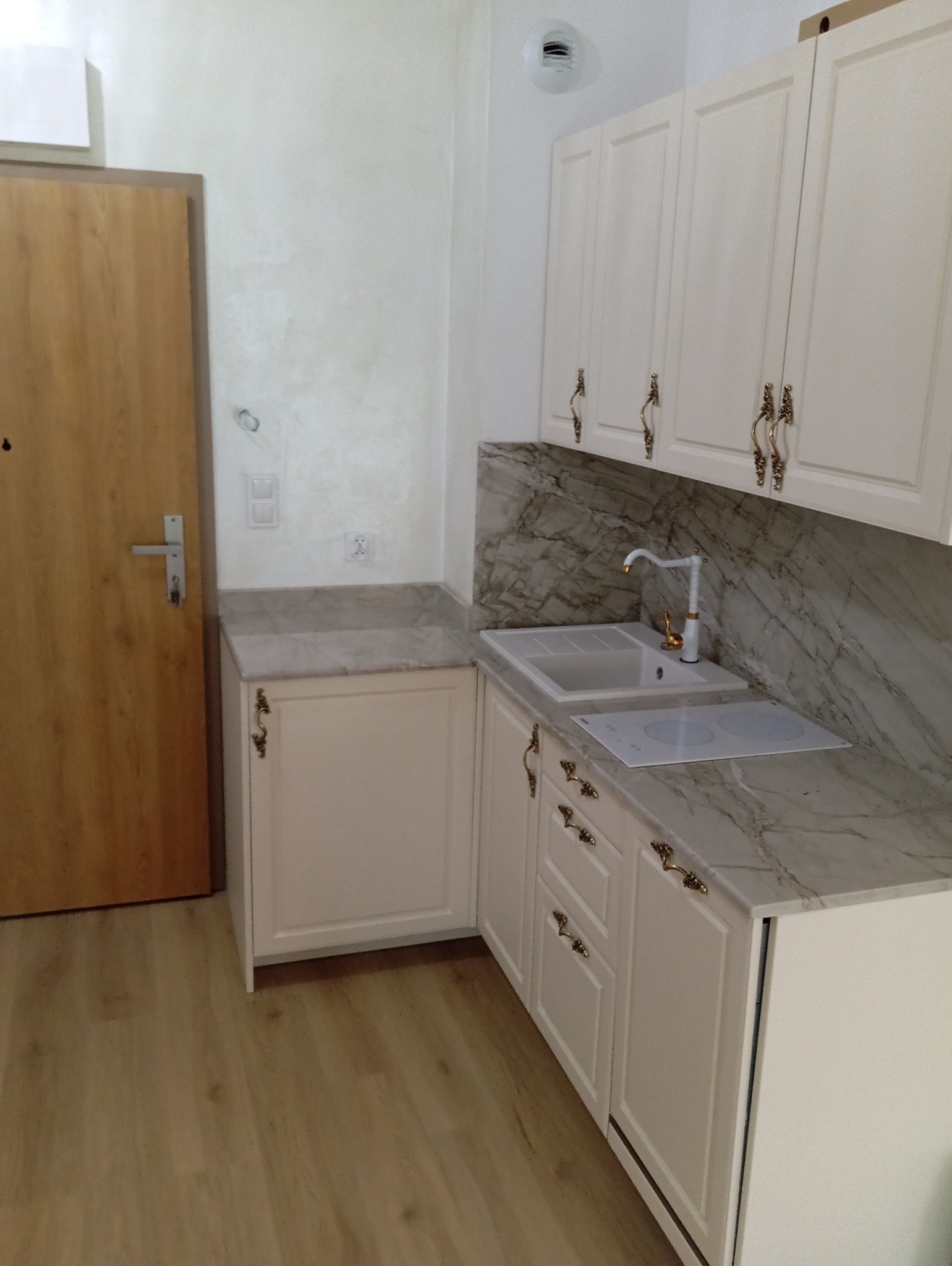 Jasna zabudowa kuchenna z marmurowym blatem i złotymi uchwytami, widoczna obok drewnianych drzwi wejściowych. Minimalistyczny design, eleganckie wykończenie.