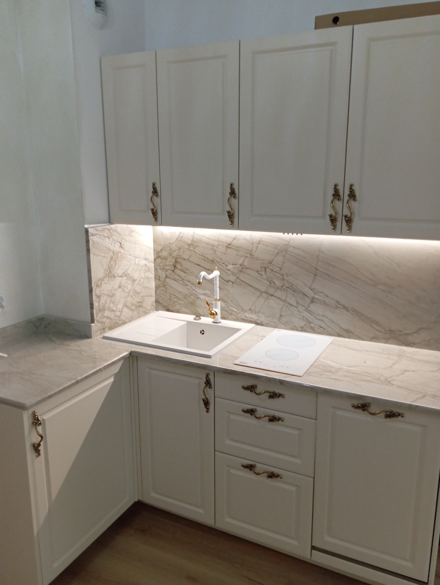 Elegancka kuchnia na wymiar w Pasieczniku z białymi frontami, marmurowym blatem i złotymi uchwytami. Widoczny zlew i płyta indukcyjna, oświetlenie LED pod szafkami.