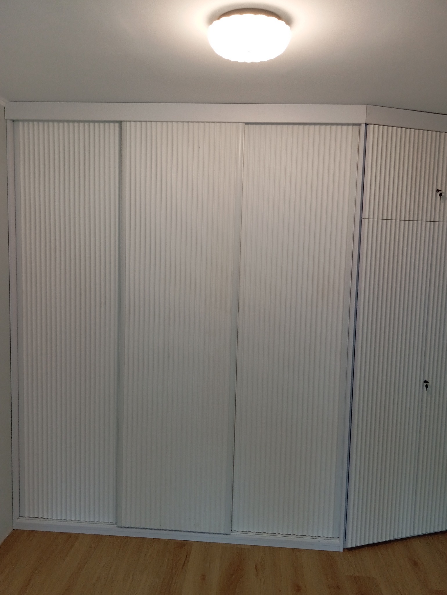 Biała szafa wnękowa z przesuwnymi drzwiami o ryflowanej fakturze, dopasowana do skosu pomieszczenia, z widoczną podłogą z paneli.