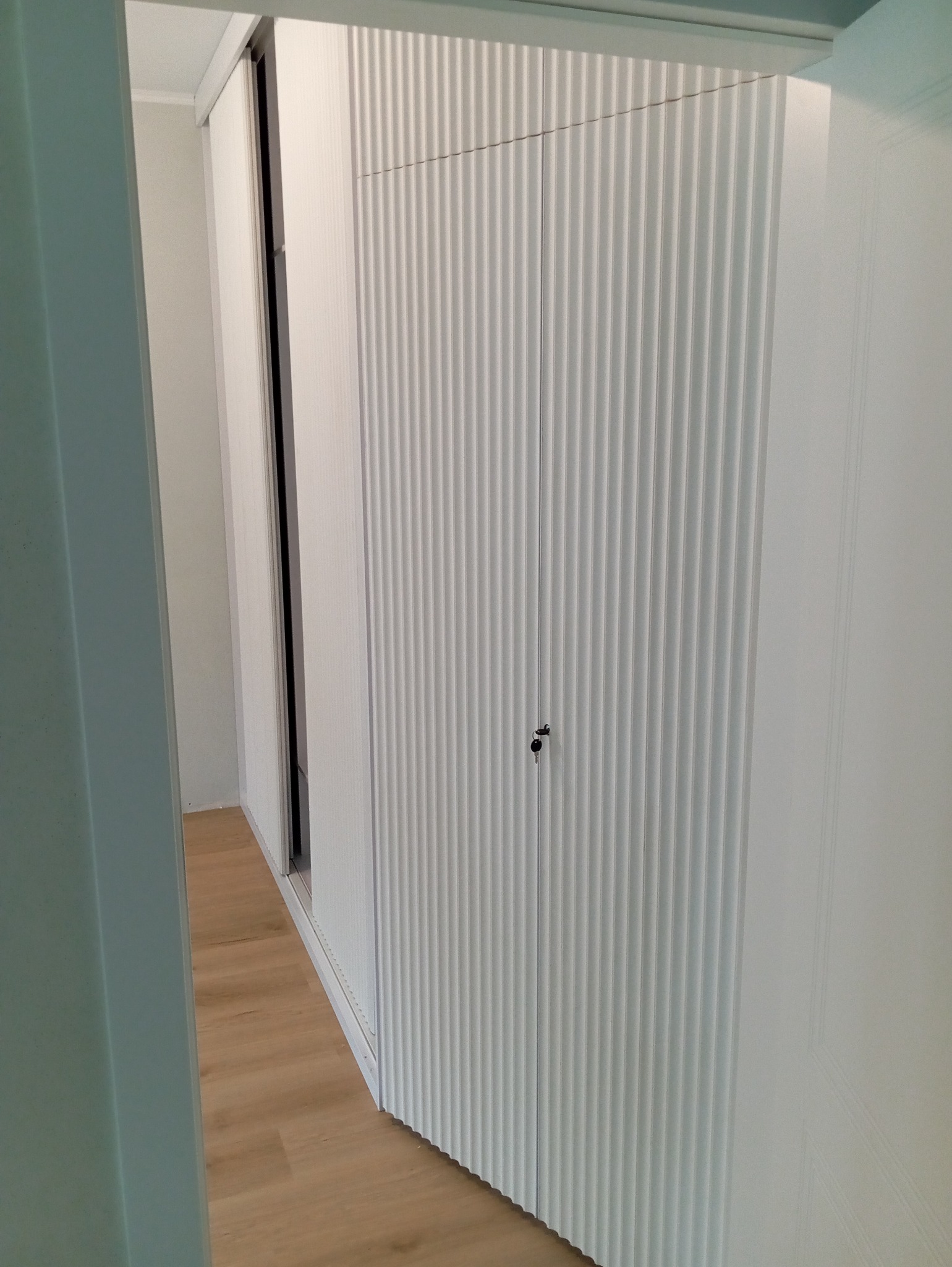 Biała szafa wnękowa z ryflowanymi drzwiami, widoczna z perspektywy wejścia do pomieszczenia, z fragmentem podłogi z paneli. Minimalistyczny design.