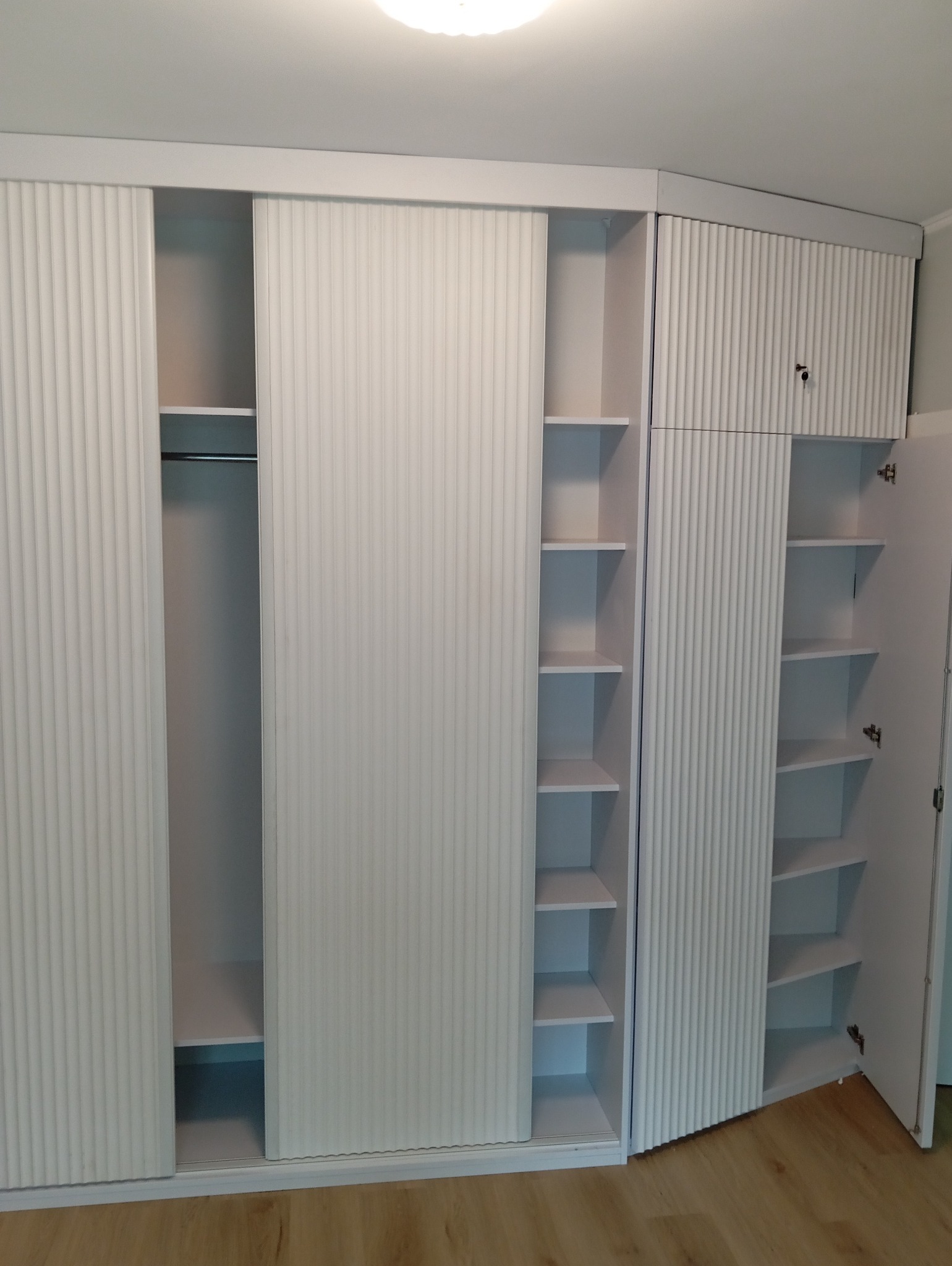 Biała szafa wnękowa z przesuwnymi drzwiami i ryflowanymi frontami. Widoczne półki i drążek na ubrania. Otwarta szafka zamykana na klucz.