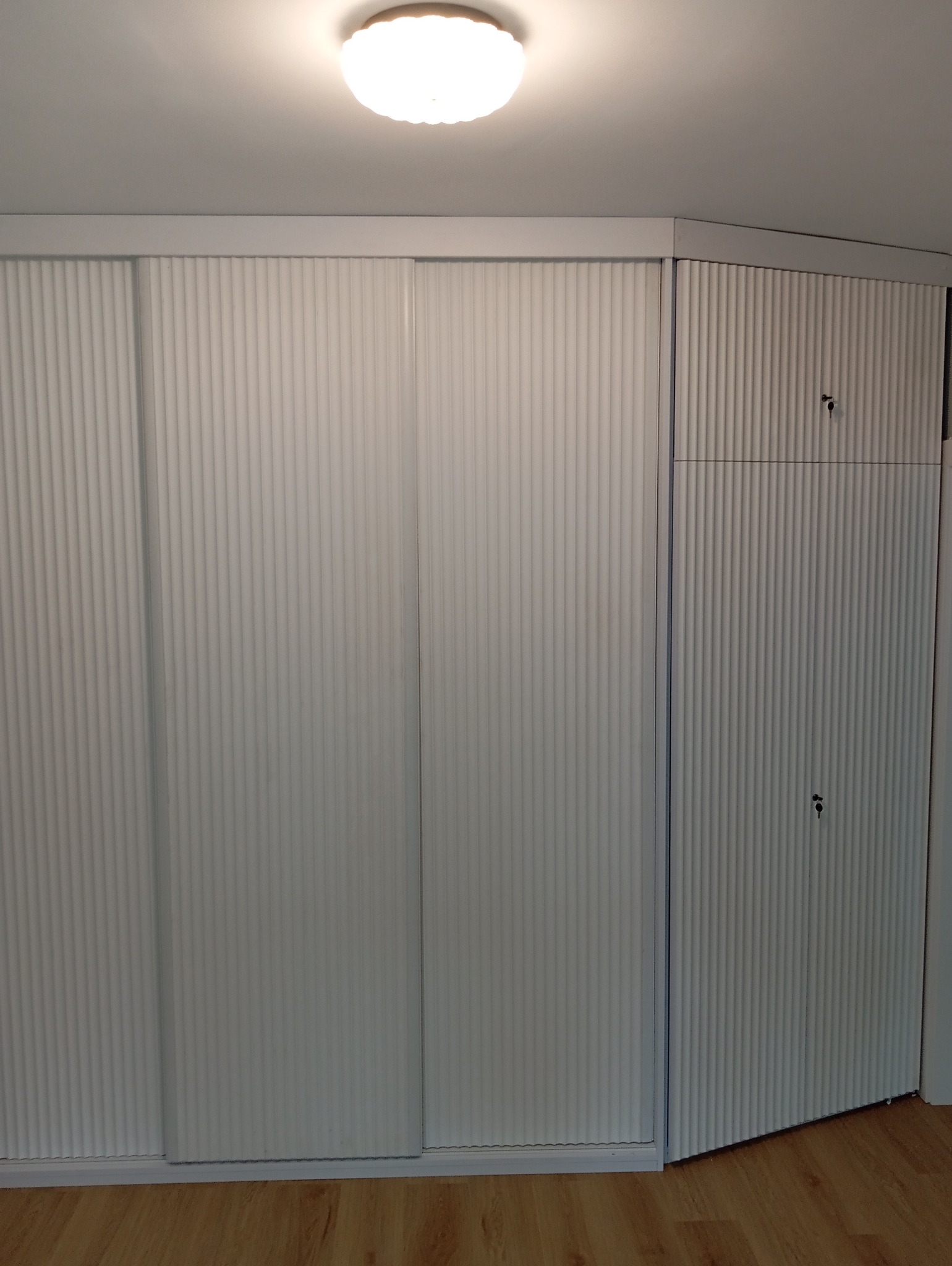 Biała szafa wnękowa z przesuwnymi drzwiami i ryflowaną fakturą, dopasowana do rogu pomieszczenia, z widocznymi zamkami w górnej części.