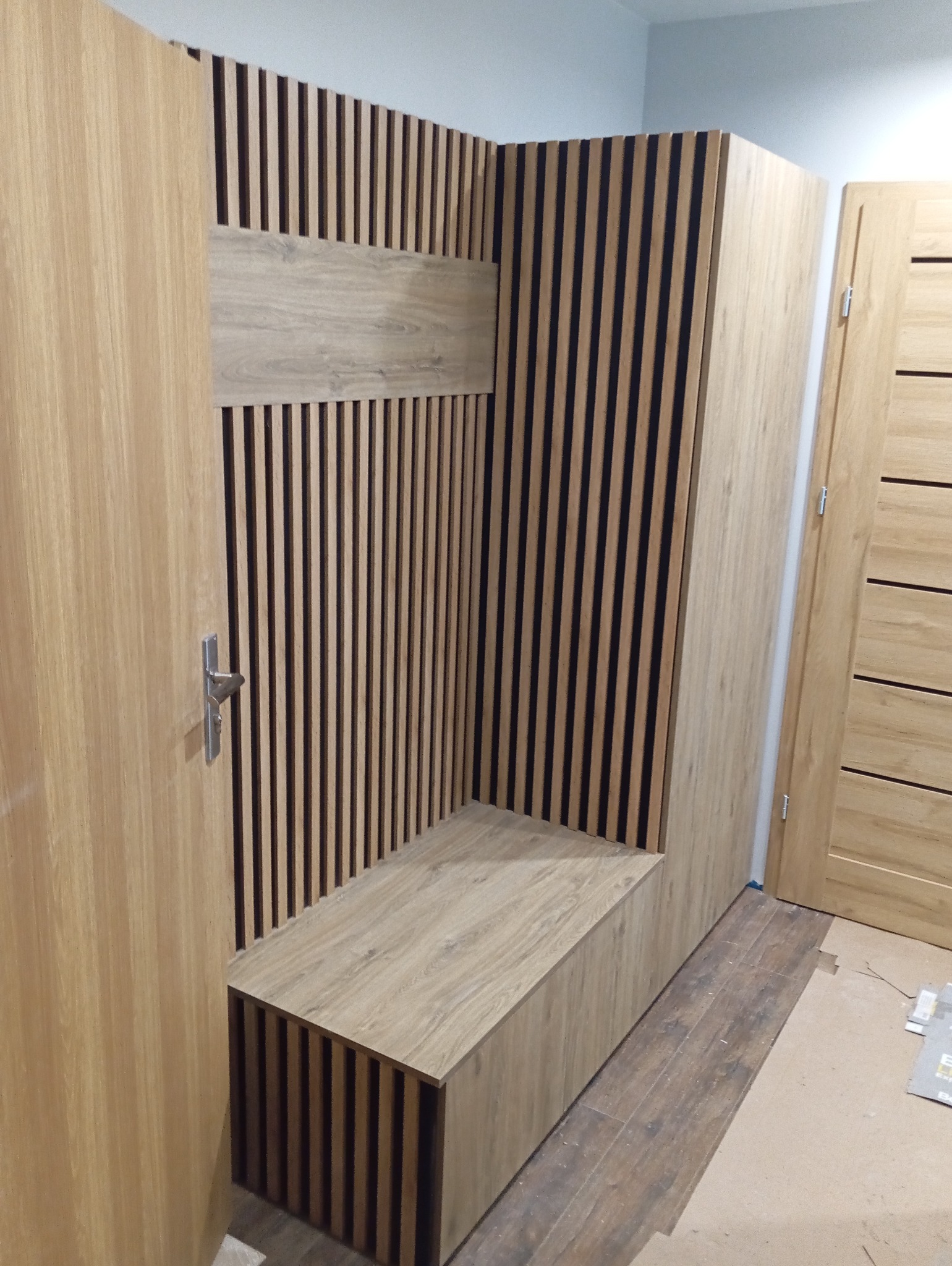 Drewniana zabudowa wnęki z siedziskiem i lamelami w odcieniach beżu i brązu. Widoczna część drzwi z klamką. Podłoga wyłożona panelami.