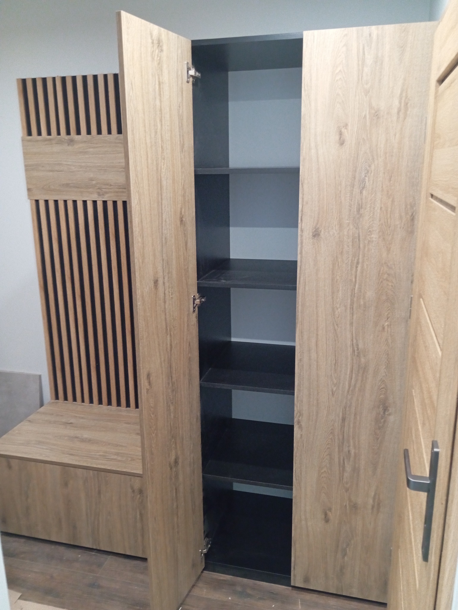 Otwarta szafa z półkami w odcieniach drewna i szarości, z elementami dekoracyjnymi na boku. Widoczne zawiasy i wnętrze szafy.