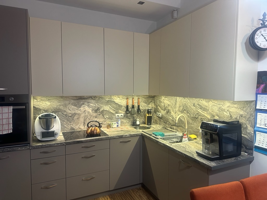 Jasna, narożna kuchnia z szarymi frontami i marmurowym blatem. Podświetlenie LED podkreśla wzór kamienia. Na blacie ekspres do kawy i akcesoria kuchenne.