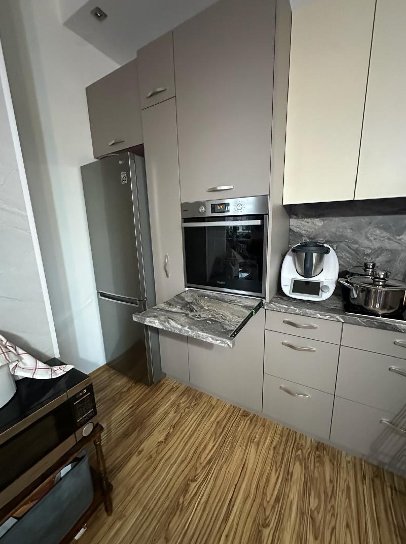 Jasna kuchnia z drewnianą podłogą, szarymi szafkami i wysuwanym blatem z marmurowym wzorem. Widoczny sprzęt AGD: lodówka, piekarnik i Thermomix.