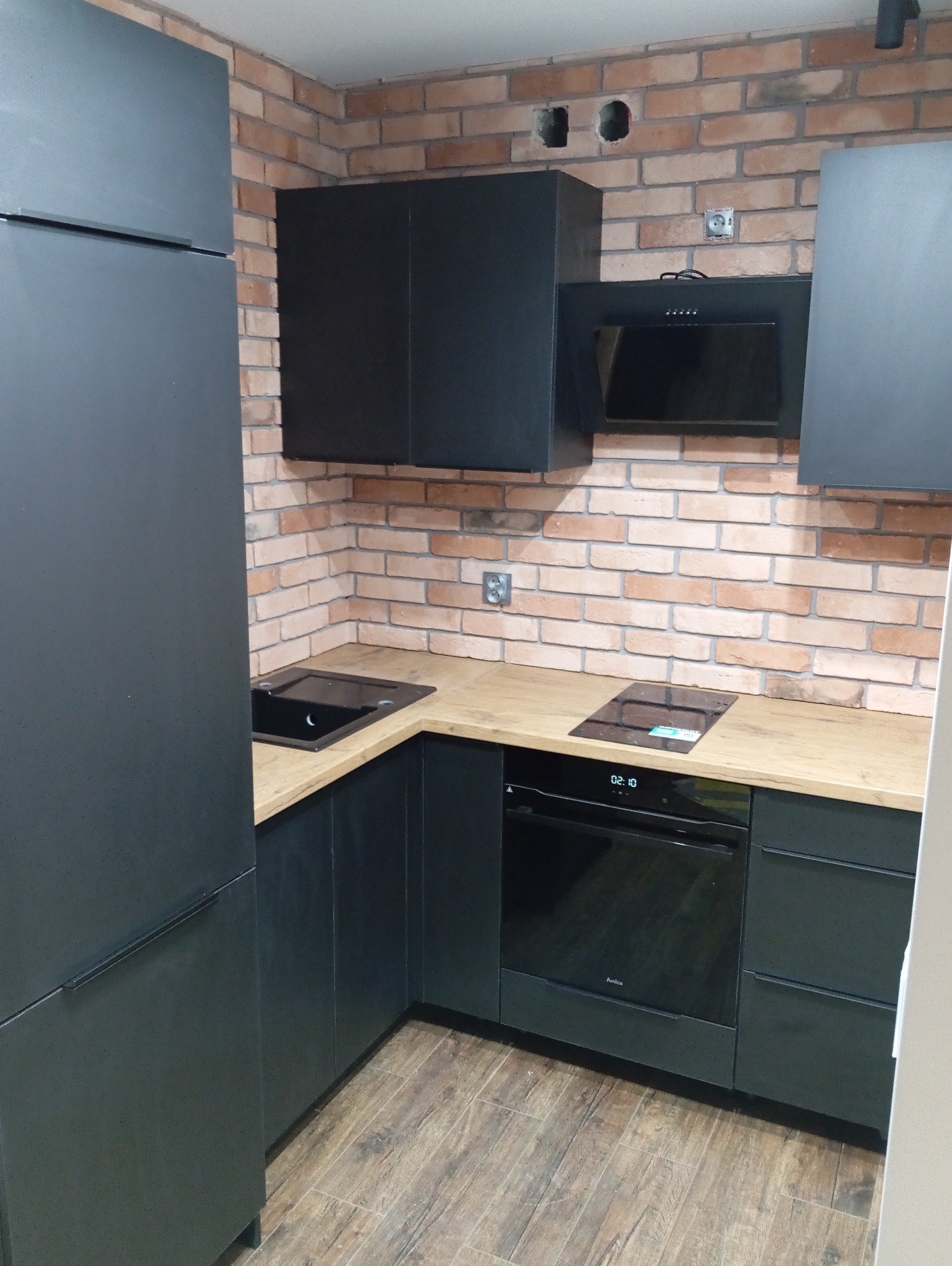 Nowoczesna kuchnia z czarnymi szafkami i blatem imitującym drewno, na tle ściany z czerwonej cegły. Widoczna płyta indukcyjna i piekarnik.