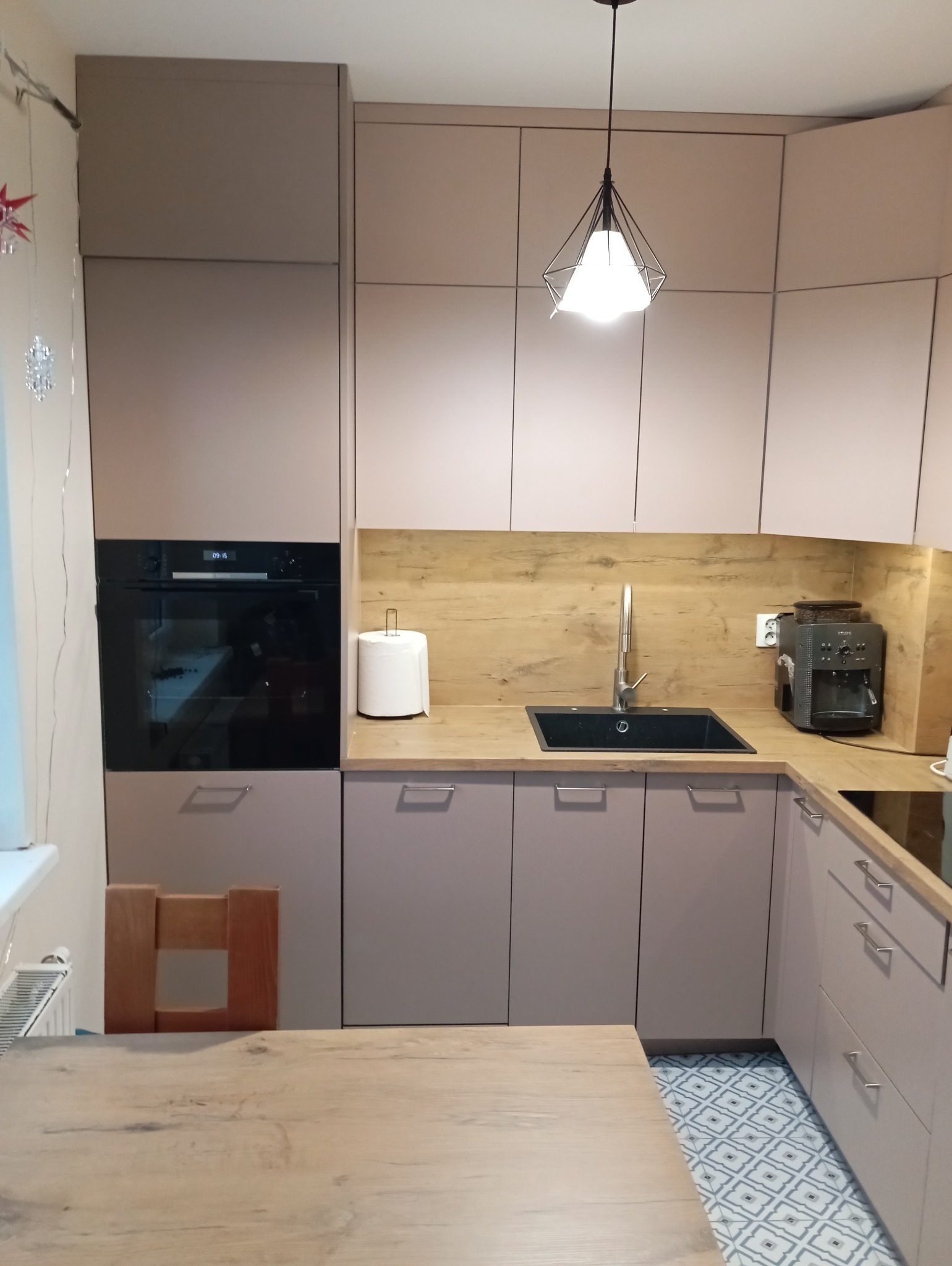 Jasna kuchnia z zabudową w odcieniach beżu i drewna. Widoczny zlew, ekspres do kawy i stół z drewnianym blatem. Nad blatem wisi nowoczesna lampa.