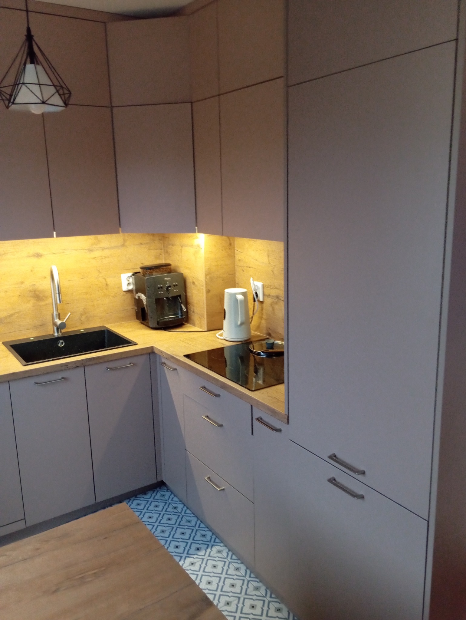 Jasnoszara zabudowa kuchenna z drewnianym blatem, czarnym zlewem i płytą indukcyjną. Na blacie ekspres do kawy i czajnik. Podłoga w niebieskie wzory.