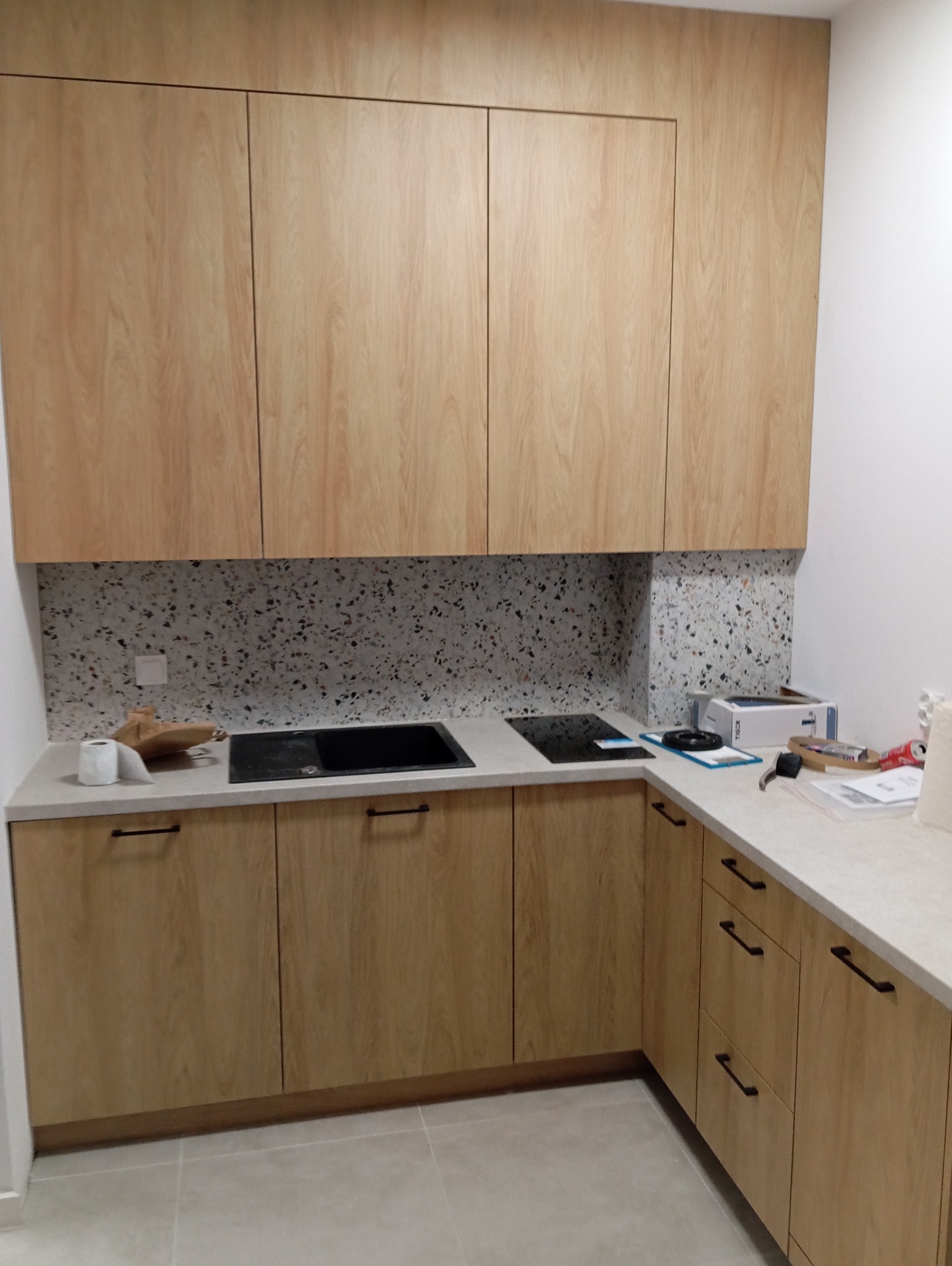 Jasna kuchnia na wymiar z drewnianymi frontami i blatem w kolorze betonu. Na blacie płyta indukcyjna. Ściana nad blatem wykończona płytkami lastryko.