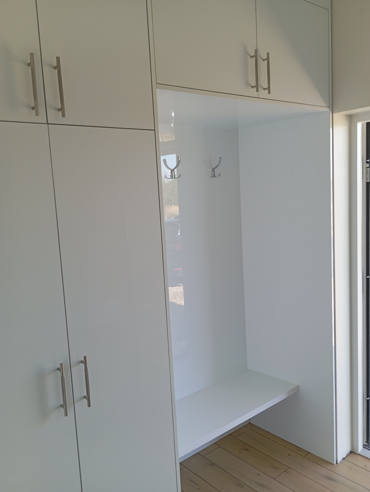 Biała szafa wnękowa z otwartą przestrzenią na wieszaki i siedzisko, widoczne metalowe uchwyty i drewniana podłoga. Minimalistyczny design, jasne wnętrze.