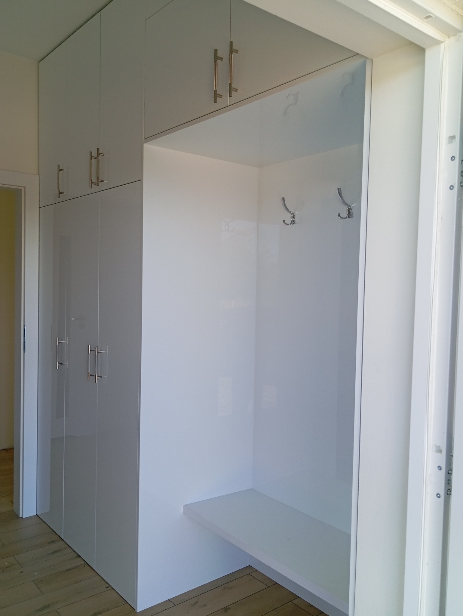 Biała szafa wnękowa z połyskiem, z siedziskiem i wieszakami, dopasowana do przedpokoju. Minimalistyczny design, metalowe uchwyty, drewniana podłoga.
