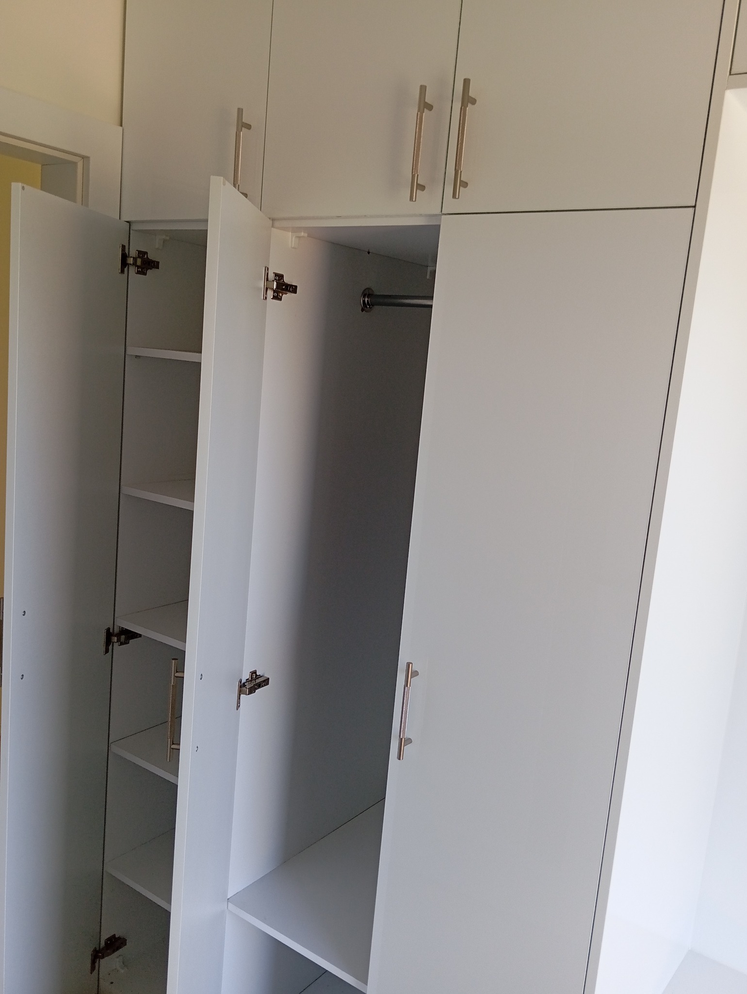 Biała szafa z otwartymi drzwiami, widoczne półki i drążek na ubrania. Minimalistyczny design, metalowe uchwyty. Szafa sięga do sufitu, jasne wnętrze.