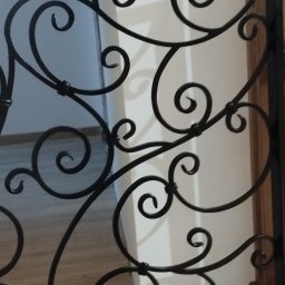 Robert Kochański - Kute, czarne zdobienie balustrady z motywem spiralnych zawijasów, widoczne z bliska, z fragmentem podłogi i ściany w tle. Dekoracyjny detal.