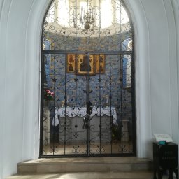 Robert Kochański - Kuta, czarna brama z dekoracyjnymi elementami, prowadząca do kaplicy z mozaiką i oświetlona żyrandolem. Widok z dołu, przez łukowate wejście.