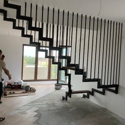 Robert Kochański - Nowoczesne schody wspornikowe z czarną, metalową konstrukcją i drewnianymi stopniami, zamontowane w jasnym wnętrzu w trakcie wykończenia. Widoczne narzędzia i materiały budowlane.