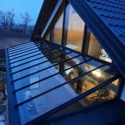 Robert Kochański - Szklana zabudowa balkonu z czarną ramą, widok z góry na konstrukcję i wnętrze. W tle pola i drzewa o zmierzchu. Nietypowy kształt dachu.