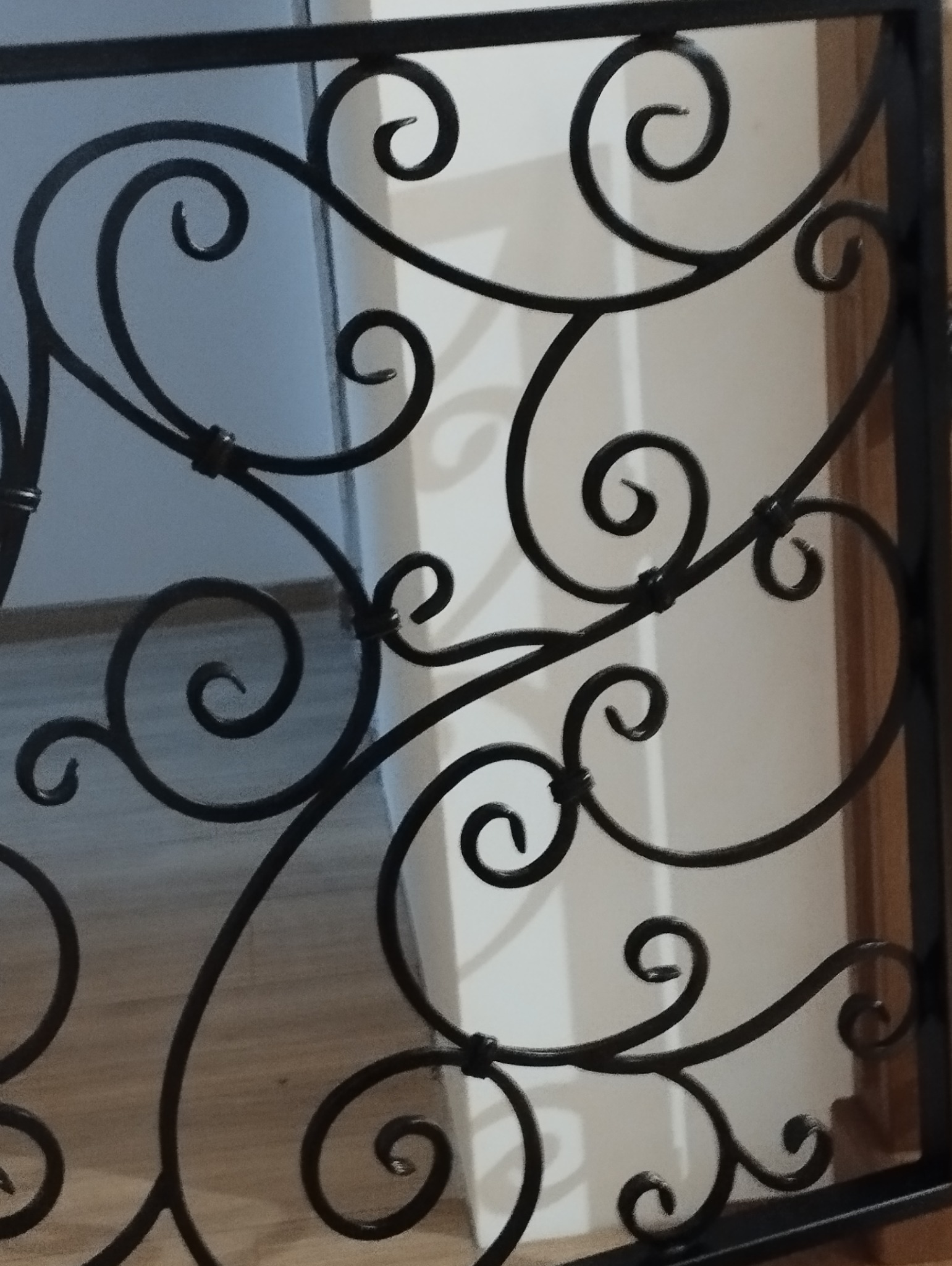 Kute, czarne zdobienie balustrady z motywem spiralnych zawijasów, widoczne z bliska, z fragmentem podłogi i ściany w tle. Dekoracyjny detal.