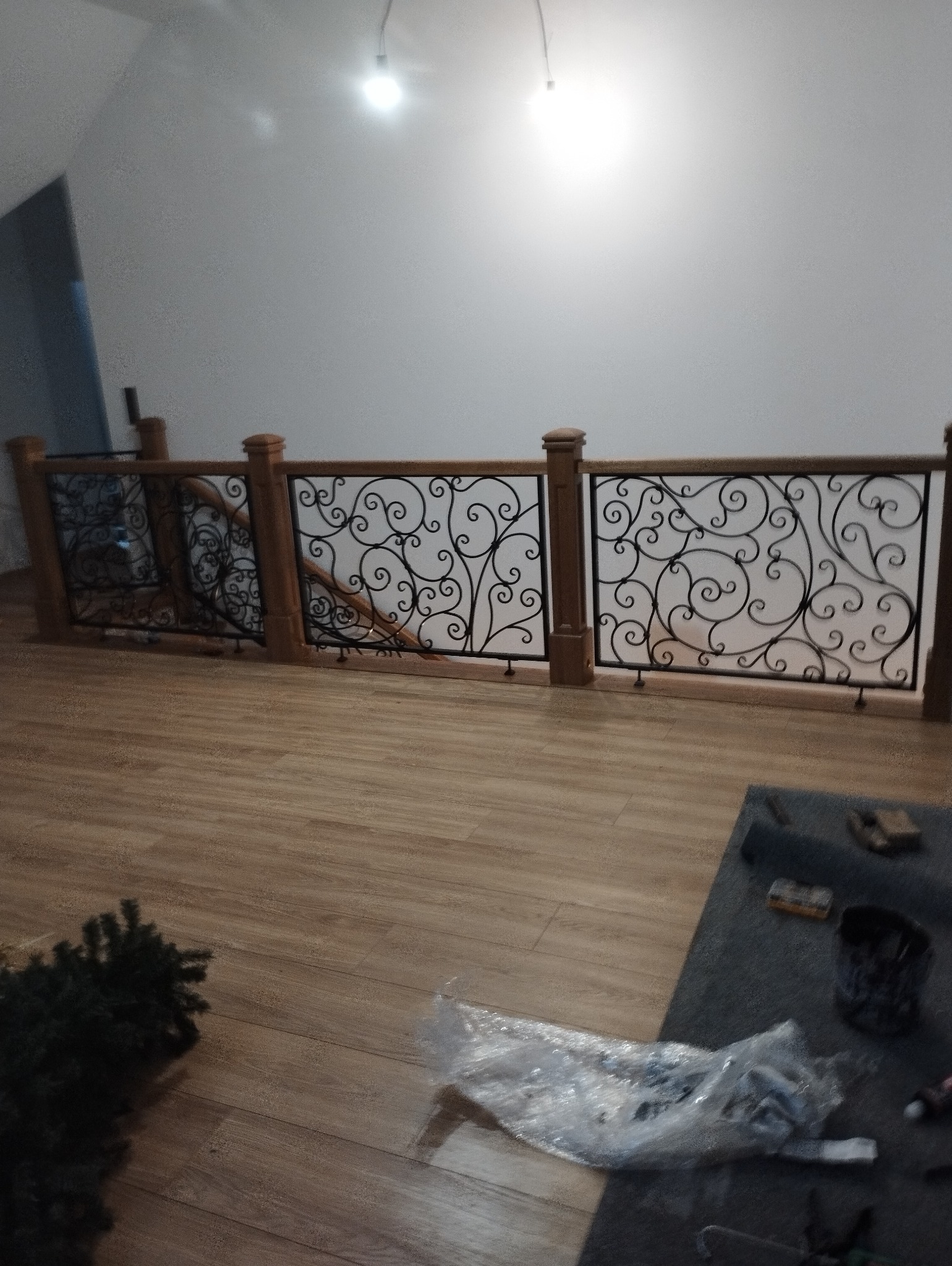 Kuta balustrada z drewnianymi słupkami, zdobiona motywem roślinnym, zamontowana wewnątrz budynku. Widoczne drewniane panele podłogowe i fragmenty narzędzi.