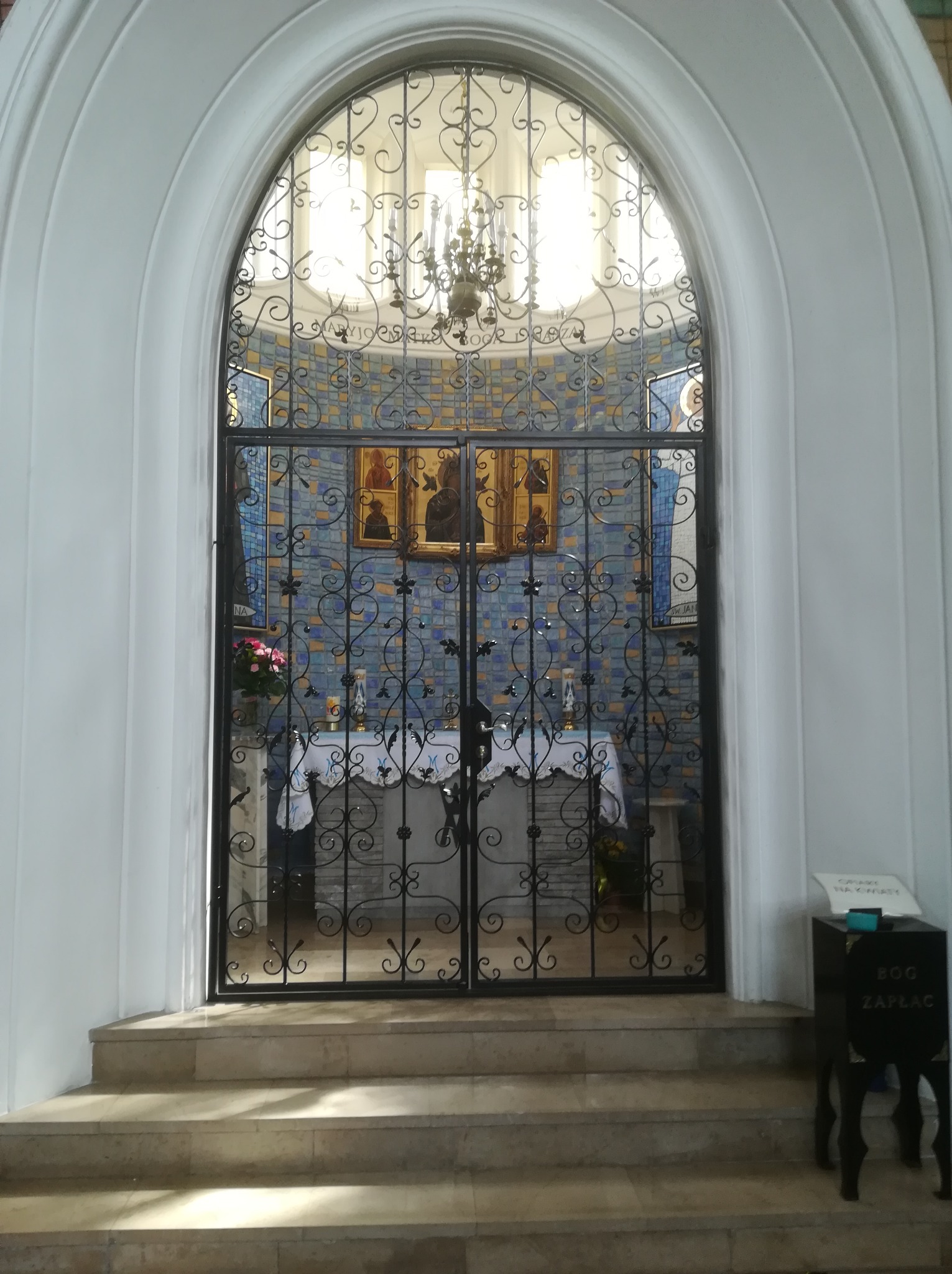 Kuta, czarna brama z dekoracyjnymi elementami, prowadząca do kaplicy z mozaiką i oświetlona żyrandolem. Widok z dołu, przez łukowate wejście.