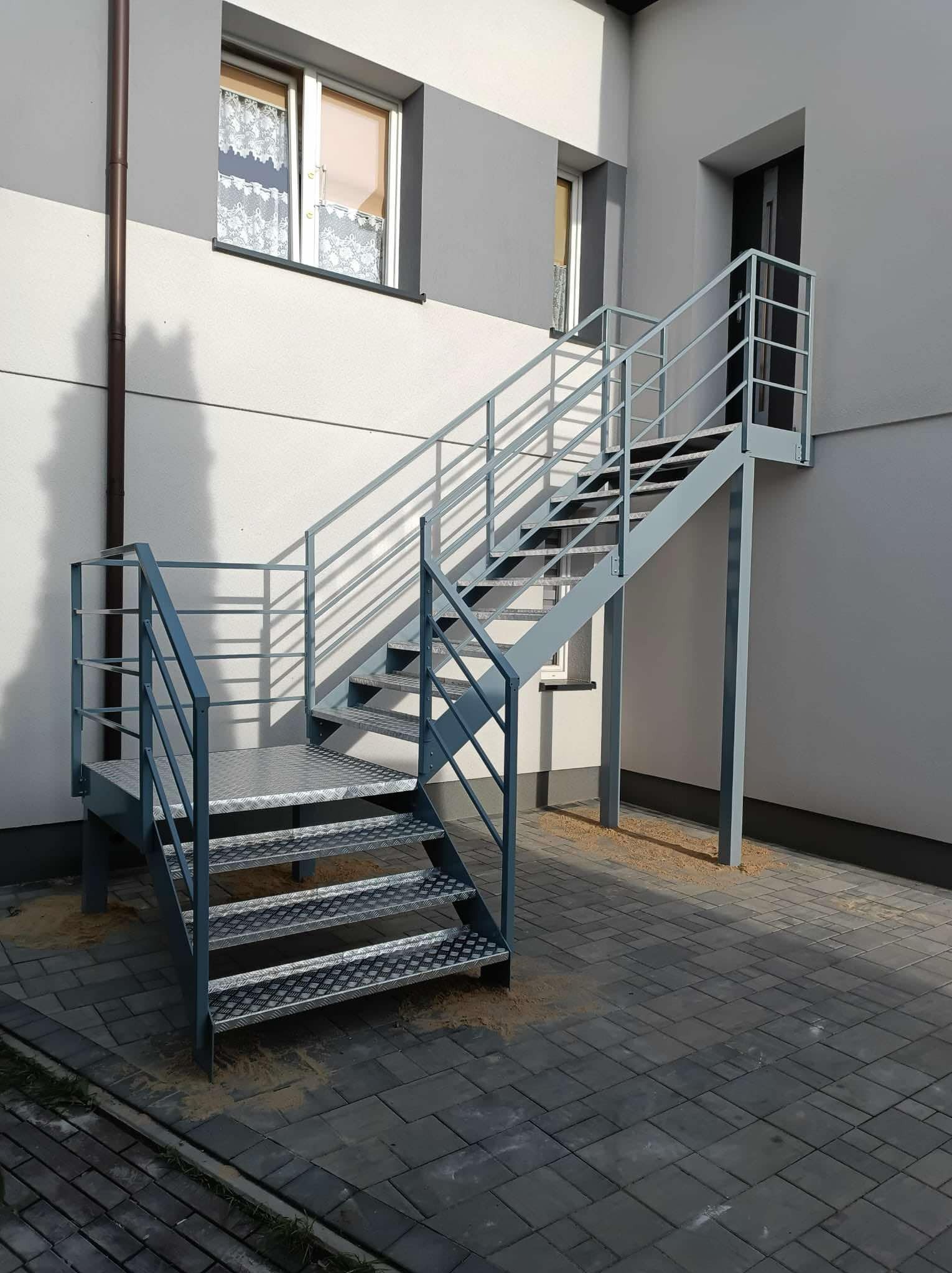 Zewnętrzne, metalowe schody z balustradą, prowadzące do drzwi na piętrze budynku. Schody w kolorze szarym, z ażurowymi stopniami. Widoczne cienie rzucane przez konstrukcję.