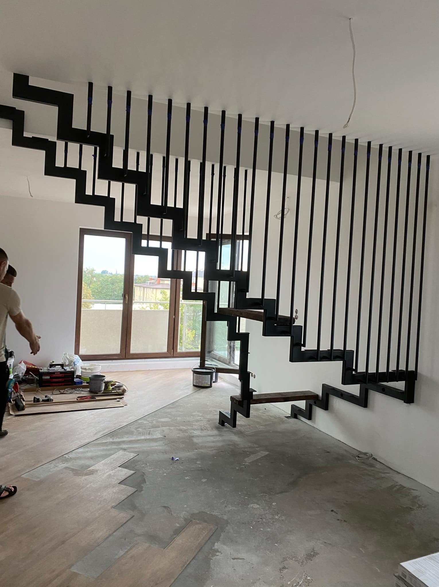 Nowoczesne schody wspornikowe z czarną, metalową konstrukcją i drewnianymi stopniami, zamontowane w jasnym wnętrzu w trakcie wykończenia. Widoczne narzędzia i materiały budowlane.