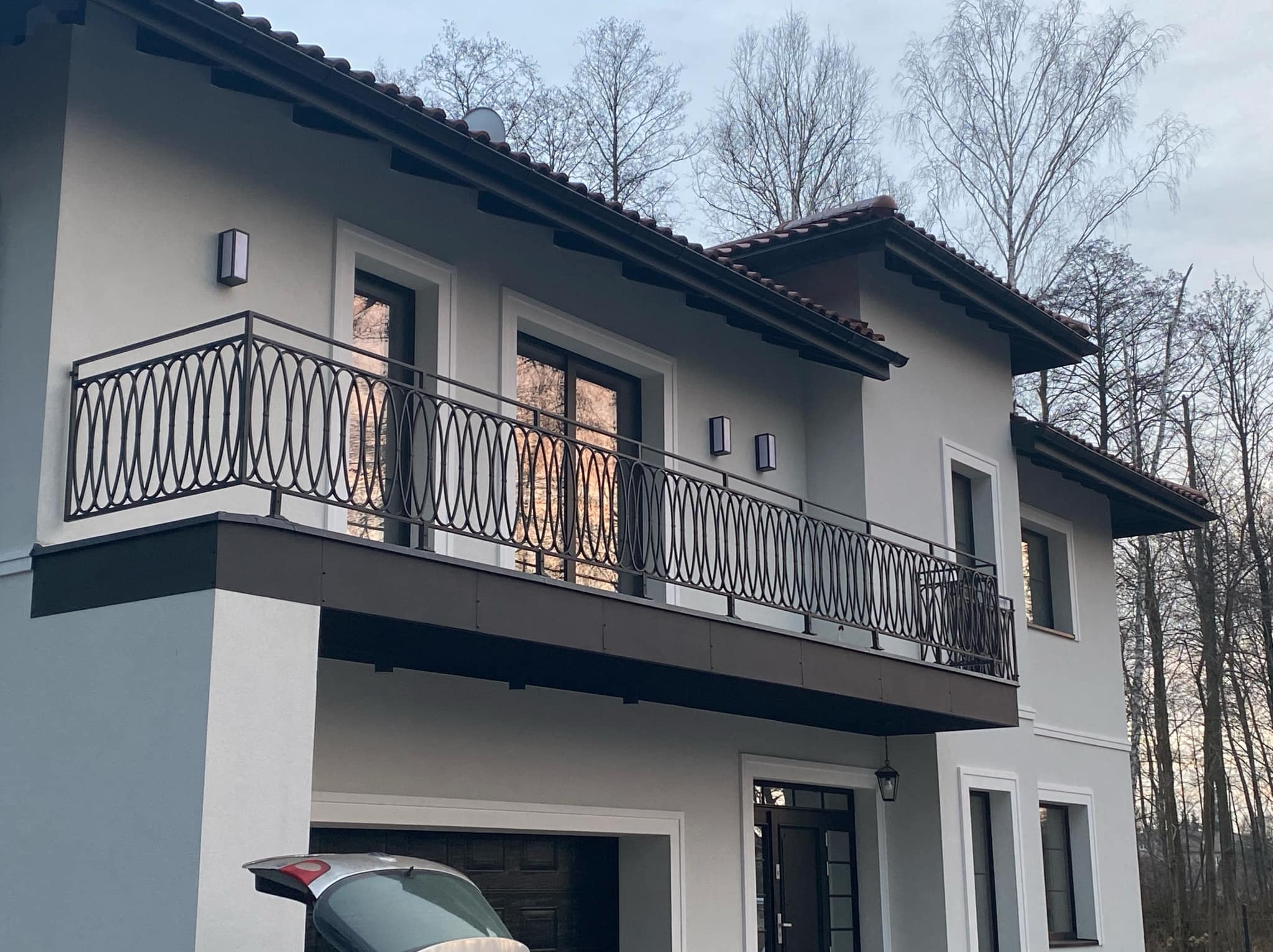 Elegancka, czarna balustrada balkonowa z dekoracyjnym wzorem na tle szarego domu z garażem. Widoczne drzewa bez liści i fragment samochodu.