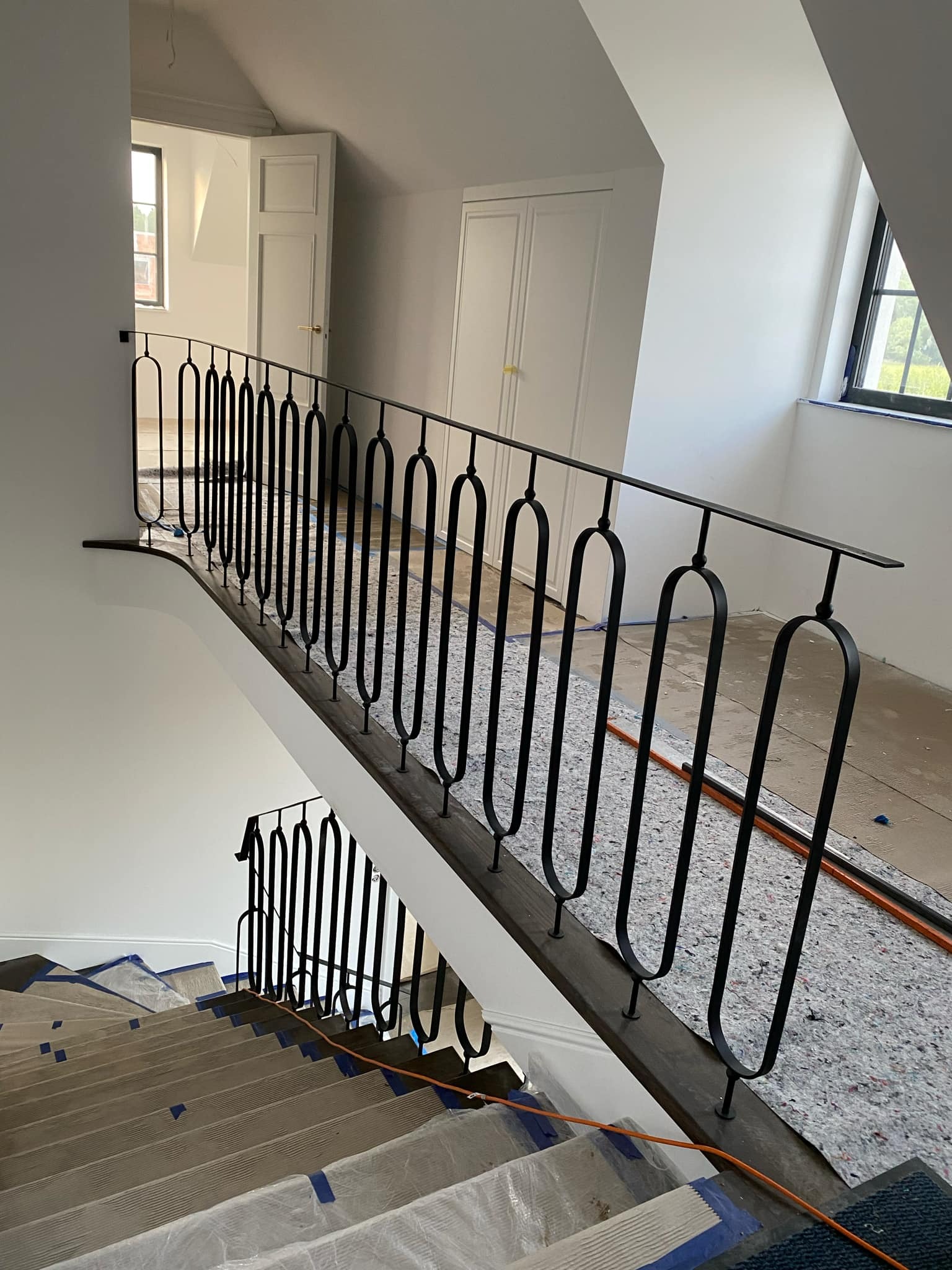 Nowoczesna, czarna balustrada schodowa z owalnymi elementami pionowymi, zamontowana w jasnym wnętrzu domu. Schody zabezpieczone folią malarską.