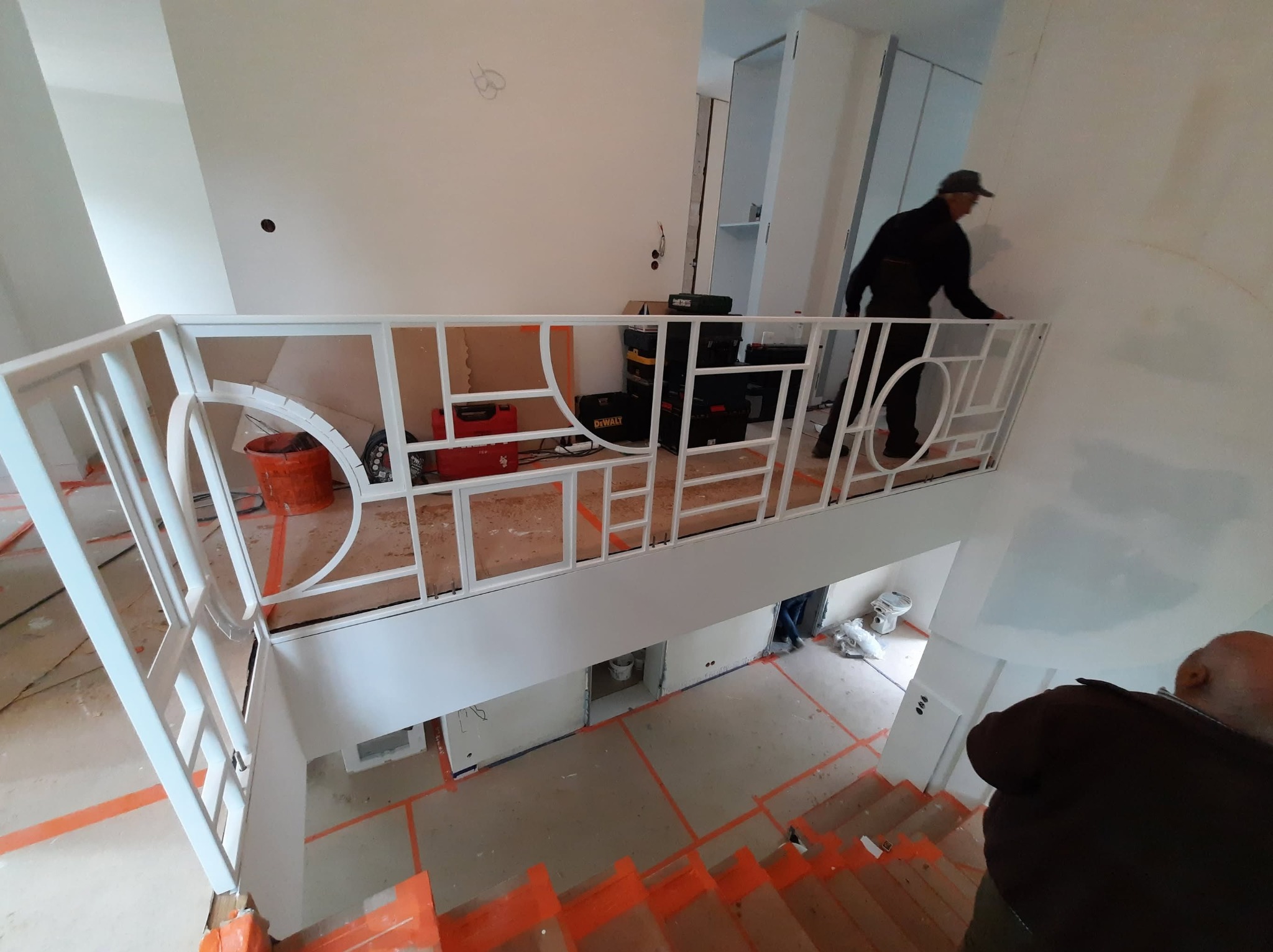 Biała balustrada z geometrycznym wzorem na antresoli w trakcie montażu. Widoczne narzędzia i osoba montująca. Pomarańczowe taśmy zabezpieczające na podłodze.