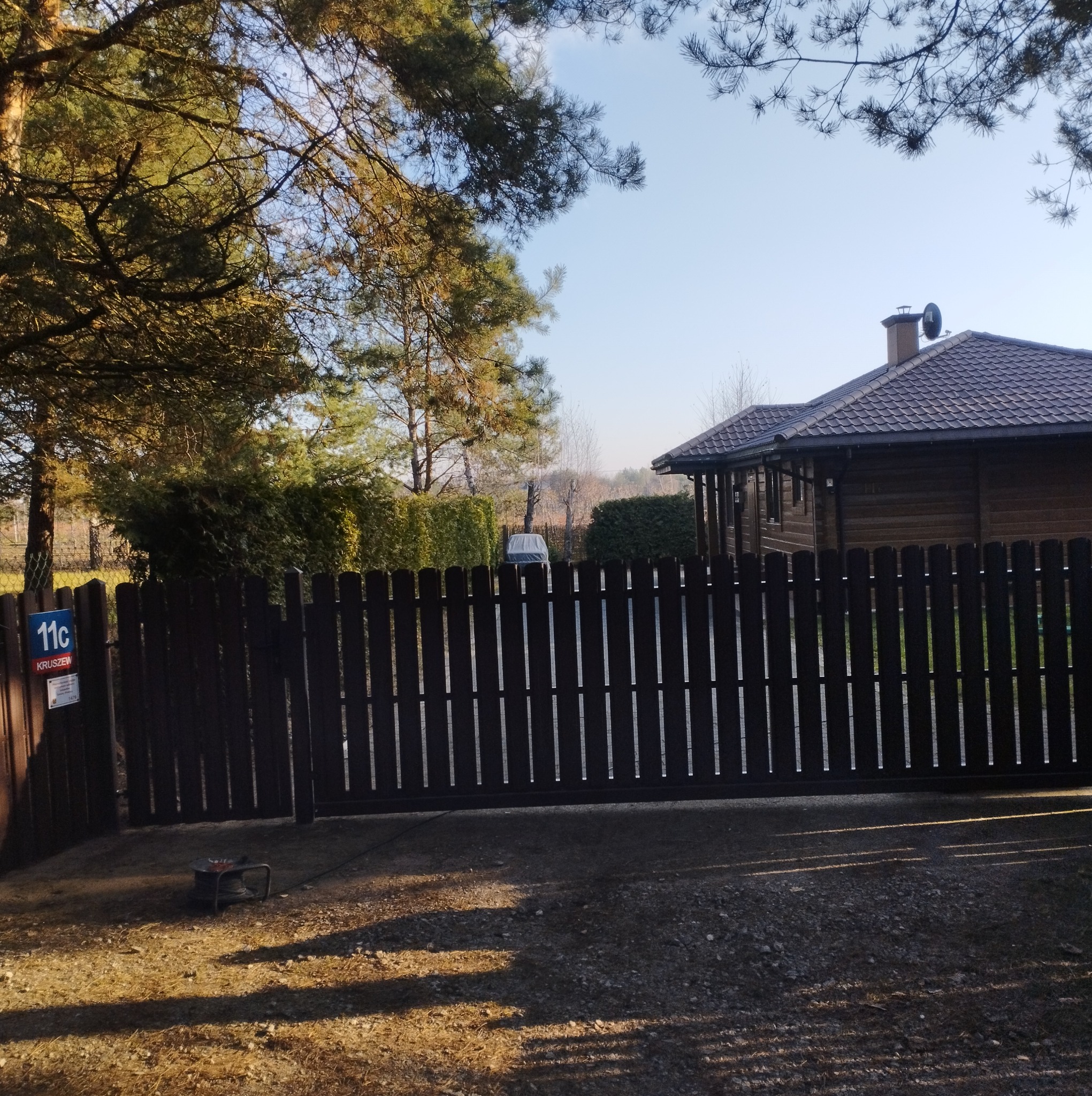 Ciemna, drewniana brama wjazdowa z pionowymi szczeblami, z numerem domu na słupku, na tle domu z brązowym dachem i zieleni drzew w słoneczny dzień.