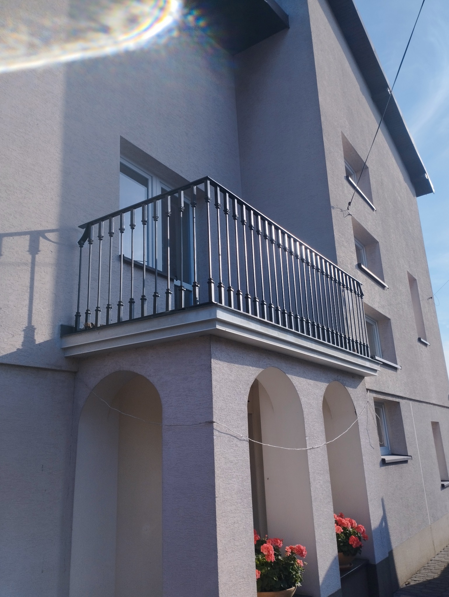 Elegancka, czarna balustrada balkonowa z pionowymi szczeblami, zamontowana na balkonie budynku z łukowatymi podcieniami i kwiatami w donicach. Jasna elewacja, błękitne niebo.